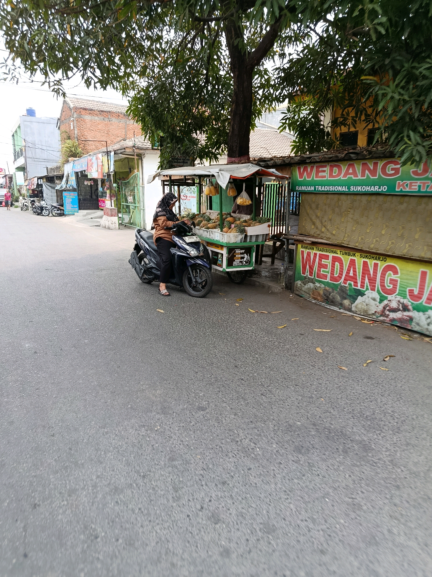 In Bekasi, Indonesia, on a paved street lined with various establishments, a woman wearing a brown jacket and black hijab sits on a dark blue motorcycle. She is positioned in front of a food stall that displays pineapples and bags of what appear to be snacks. The stall is adorned with colorful banners, one of which reads "WEDANG JA" in large red letters, and another smaller banner beneath it states "RAMUAN TRADISIONAL SUKOHARJO". Behind the stall, a building with a yellow facade is visible, partially obscured by tree branches. Further down the street, other motorcycles are parked, and several low-rise buildings with red-tiled roofs and brickwork are present. The overall scene depicts a typical street view in an Indonesian city, with a focus on the food vendor and the immediate surroundings.