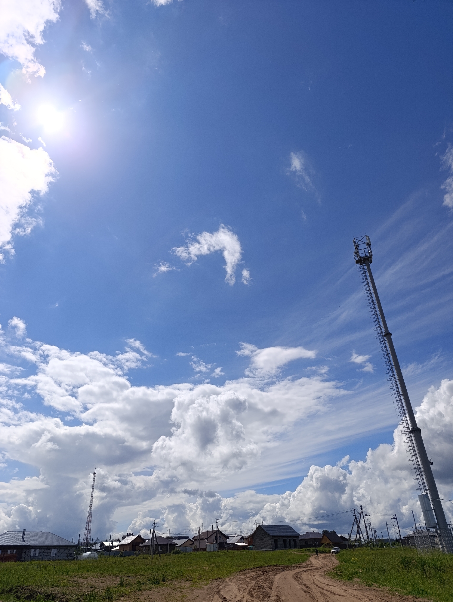 The image captures a rural scene in Chaikovsky, Russia, under a bright, partly cloudy sky with the sun visible at the top left. A dirt road leads into the scene, bordered by green grass and vegetation. A cluster of houses lines the horizon, along with utility poles and power lines. In the middle ground, two cell towers stand prominently. The larger tower on the right has a ladder extending to the top. The foreground shows wheel tracks in the dirt road. A light-colored car is visible in the distance between the houses. The overall impression is a sunny summer day in a village or outskirts area near Chaikovsky.