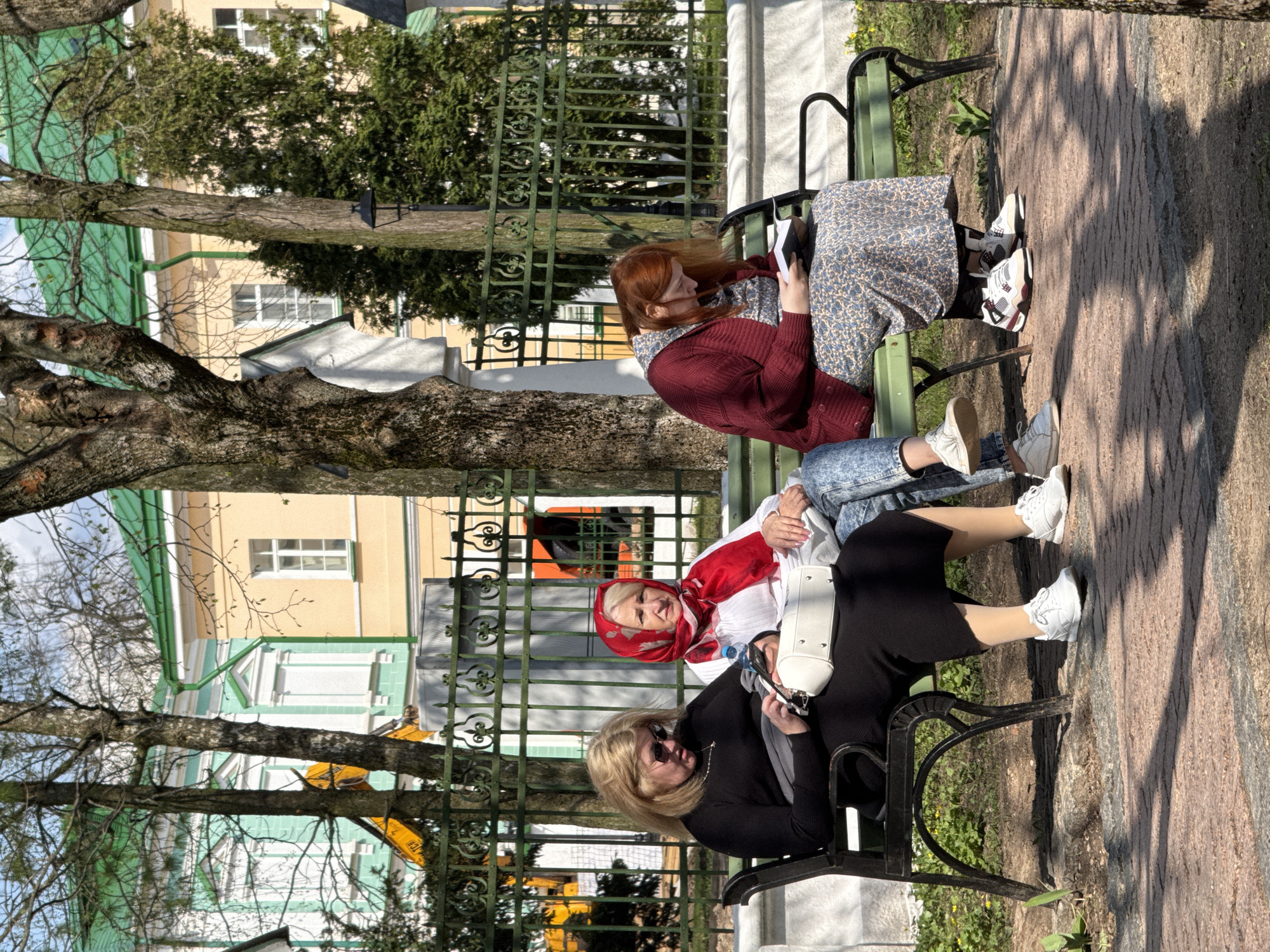 Three women are seated on a green bench in an outdoor setting. One woman has red hair, is wearing a maroon cardigan and a printed dress, and is engrossed in reading a book. Another woman in the middle is wearing a white shirt, jeans, and a red headscarf. She is looking directly at the camera. The third woman, wearing a black dress and sunglasses, is looking at a mobile phone in her hands. The bench is positioned on a brick-paved area, and behind it, there is a green iron fence. Beyond the fence, various buildings with different colors (yellow, green, white) and architectural styles are visible. Trees and branches partially obscure the view of the buildings. A construction vehicle is visible in the background. The location is the city of Unknown, Unknown.