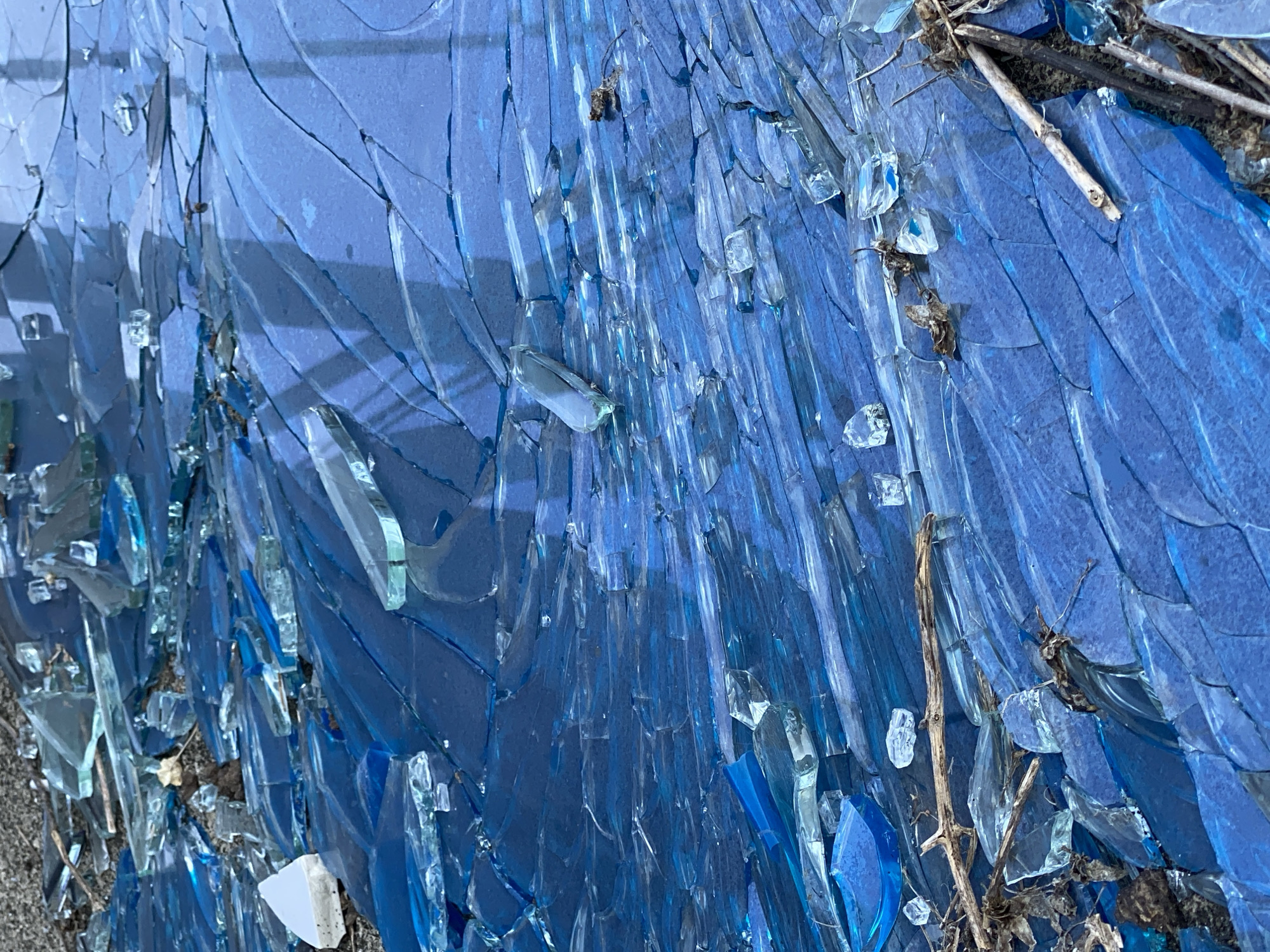 The image shows a close-up, overhead view of a pile of shattered blue glass. The glass pieces are sharp and jagged, with many different sizes and shapes. Some of the pieces are thin and flat, while others are thicker and more irregular. The glass is predominantly a vibrant blue, with some lighter shades and clear fragments visible. Shadows from an unseen overhead structure create patterns on the surface of the glass. Scattered amongst the glass are small pieces of debris, including dirt, dry leaves, and twigs. The scene appears to be outdoors, possibly on a road or a ground surface. There are no people or animals visible in the image. The overall impression is one of damage and disarray. The location in Bac Nha Trang Ward, Vietnam, is not visually evident from the provided image.