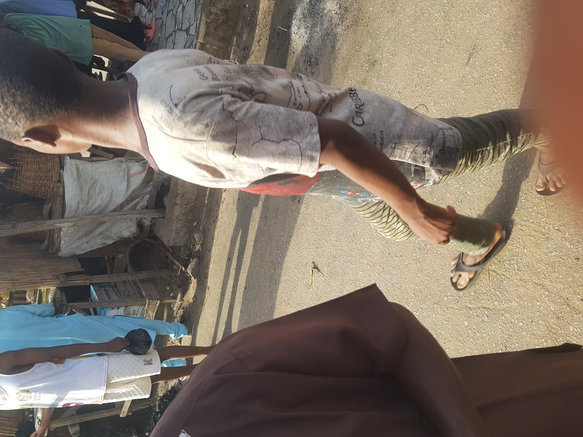 A person walks on a dirt street in Dabawa, Nigeria. The person wears sandals, striped pants, and a patterned t-shirt. Other people are nearby. Buildings of wood and metal are in the background. A brown cloth covers the lower part of the foreground.