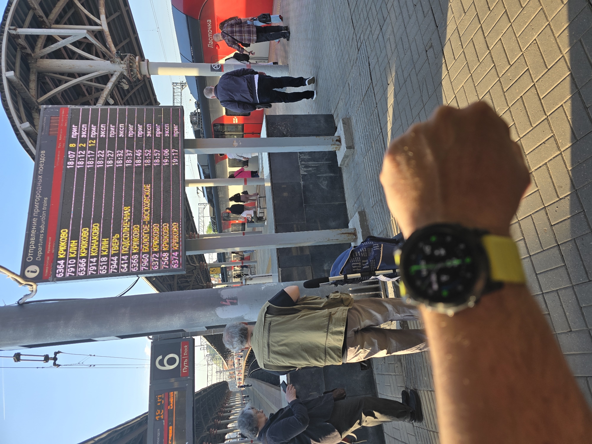 The image captures a scene at a train station in Moscow, Russia. The time indicated on a smart watch, worn on the forearm of a person, is visible in the foreground.
Several individuals are present in the scene: some waiting on the platform. A train labeled "Lastochka" (Ласточка) is parked at the station.
A departure board displays train information, including destinations such as "Kriukovo," "Klin," and "Konakovo," as well as departure times and train categories. Another sign shows that the track is number 6. The platform is paved with interlocking blocks.