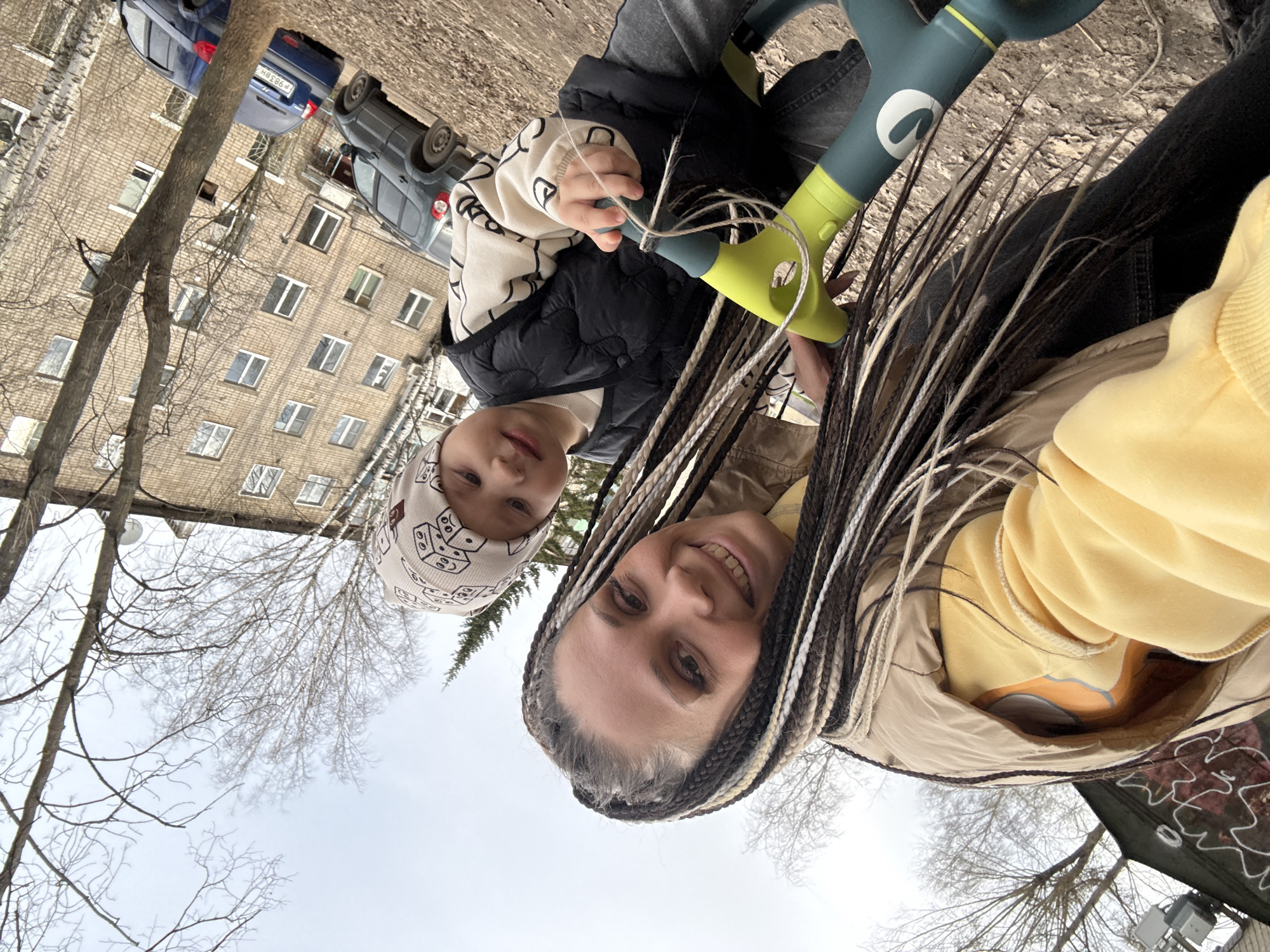 A woman and a young child are outdoors, likely in a park or residential area in Rusanovo, Russia. The woman has braided hair and is wearing a yellow sweatshirt and a light-colored jacket. The child wears a vest and a patterned hat, and is holding the handlebar of a green and black children's scooter. The background shows an apartment building, bare trees, cars, and a cloudy sky.