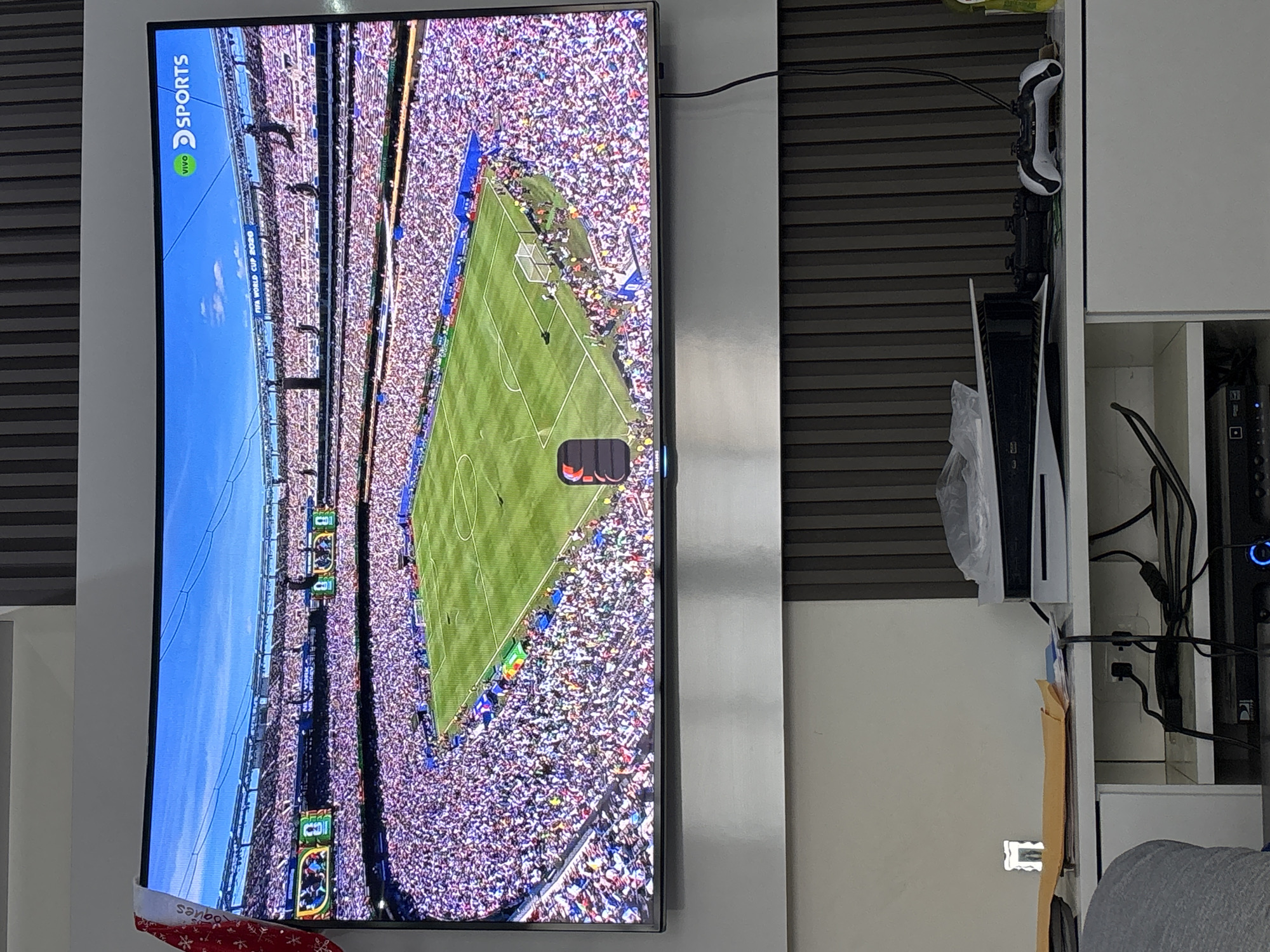 The image shows a large, curved television screen displaying a wide-angle shot of a packed soccer stadium from a high vantage point. The stadium is filled with spectators wearing predominantly white clothing, creating a sea of white in the stands. The soccer field below is green, with visible white markings and a goal at one end. Two large video screens are visible on the stadium's upper level, showing images of players. The broadcast logo "VIVO SPORTS" is in the upper right corner of the screen, along with the text "FIFA WORLD CUP 2020". A red and white festive hat, possibly a Santa hat, is draped over the top left edge of the television.
Below the television screen, a white entertainment unit is visible. On top of this unit, there is a white gaming console (likely a PlayStation 5) with a clear plastic bag covering part of it. Next to the console, two black game controllers are present, one white and black and the other black. Cables are connected to the console and power outlets. To the left of the entertainment unit, a yellow envelope is on a shelf, and a piece of a person's grey clothing is visible in the lower left corner of the frame. The background behind the entertainment unit consists of vertical wooden slats. The overall scene appears to be an indoor living room or entertainment area.