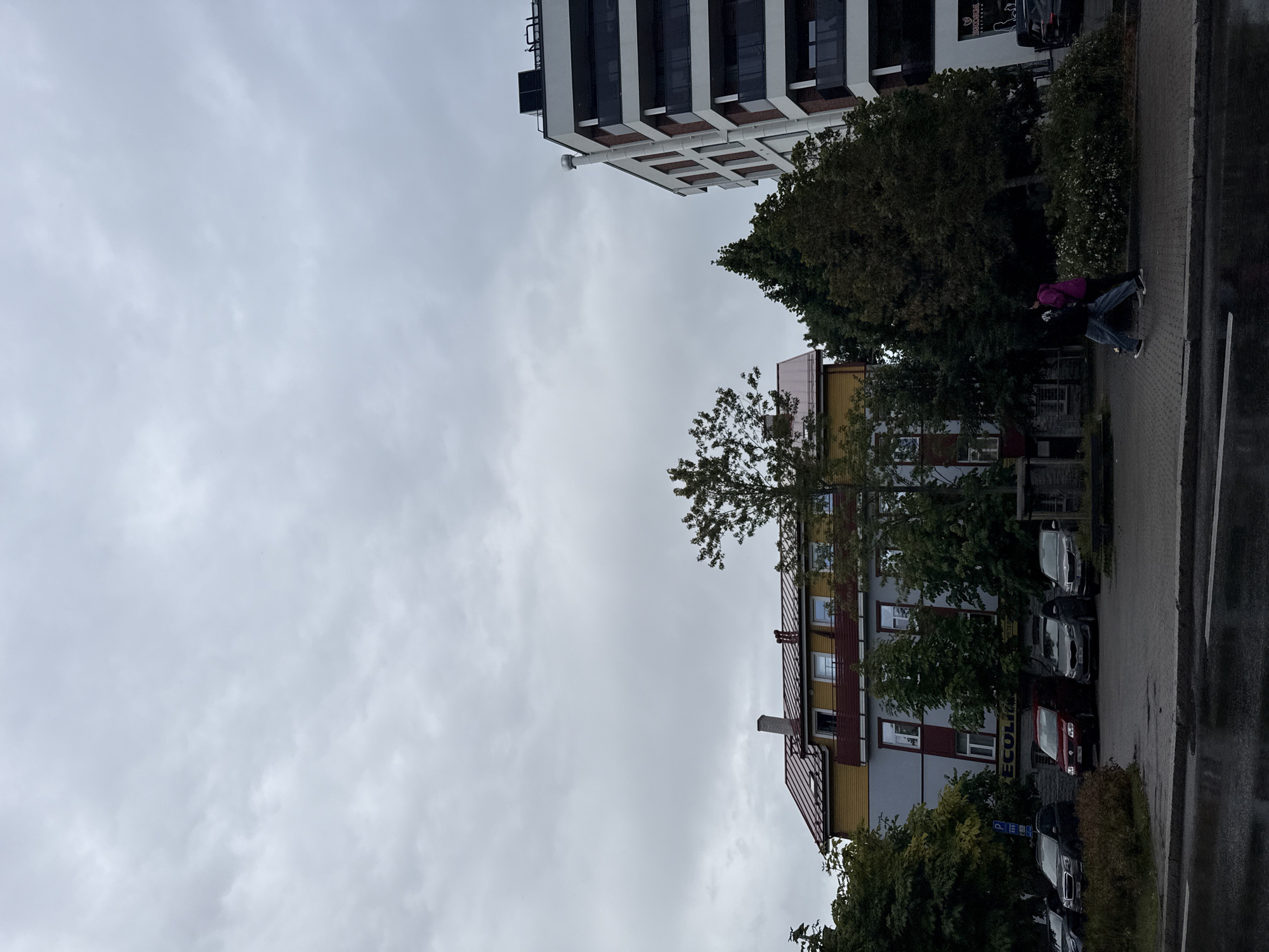 This is a photograph taken on a cloudy day in Tallinn, Estonia. The scene features a street with parked cars and a sidewalk, leading up to several buildings. On the left, a multi-story building with a yellow facade and a red roof is visible. To the right of it, a modern, white and dark-paneled apartment building stands tall. In the middle ground, a group of trees are present, separating the two main buildings. Two individuals, one wearing a pink jacket and the other in dark clothing, are walking on the sidewalk from right to left. The ground in the foreground appears wet, suggesting recent rain. The sky is overcast with gray clouds.