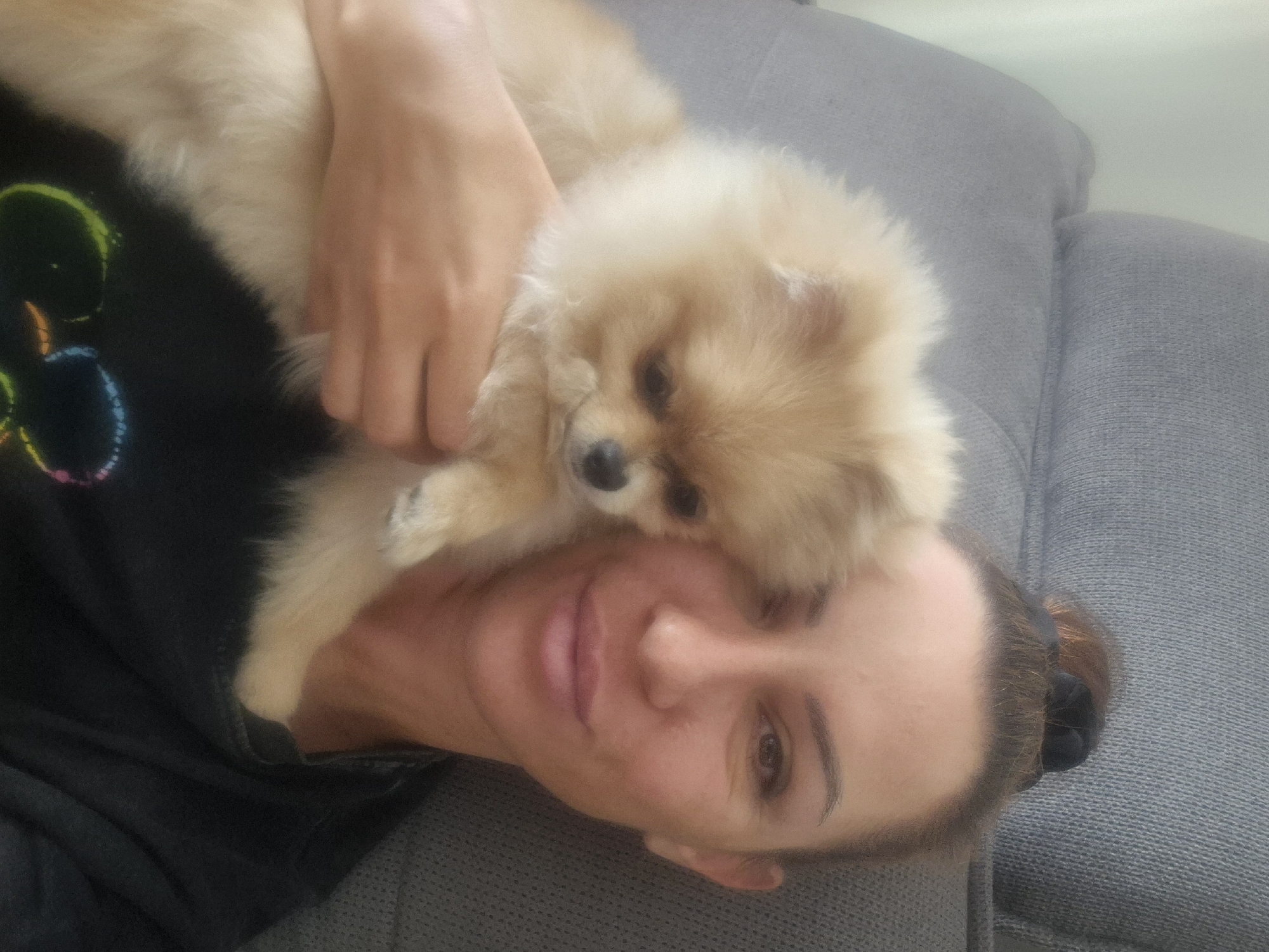 A light-skinned woman with her hair pulled up in a bun is taking a selfie while holding a light-brown Pomeranian dog. She is wearing a black t-shirt with a colorful graphic design on it. The woman and the dog are sitting on a gray sofa. The background is blurry and consists of the sofa and a wall.