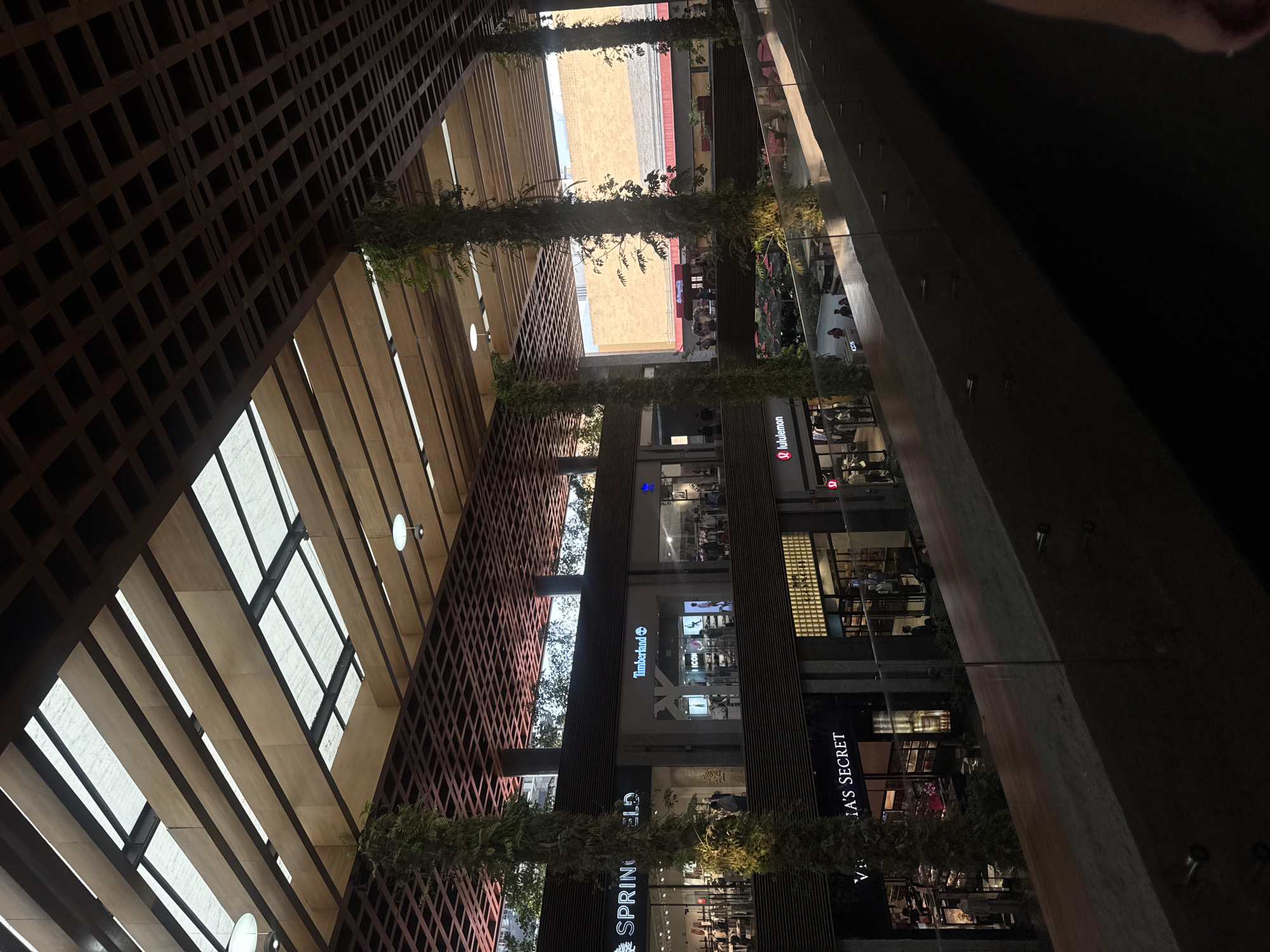 The image shows the interior of the Angelópolis Lifestyle Center in Puebla, Mexico, looking up towards the ceiling. The ceiling is constructed with a grid-like wooden structure and skylights. Columns decorated with greenery vertically bisect the open space, with retail storefronts visible on either side. Retailers with recognizable signage are Lululemon, Timberland, Springfield, and Victoria's Secret. People are seen moving within the shops. The image is taken from a low angle, emphasizing the height of the building. Part of a glass railing and a dark blurred object, possibly a hand, are visible in the upper right corner.