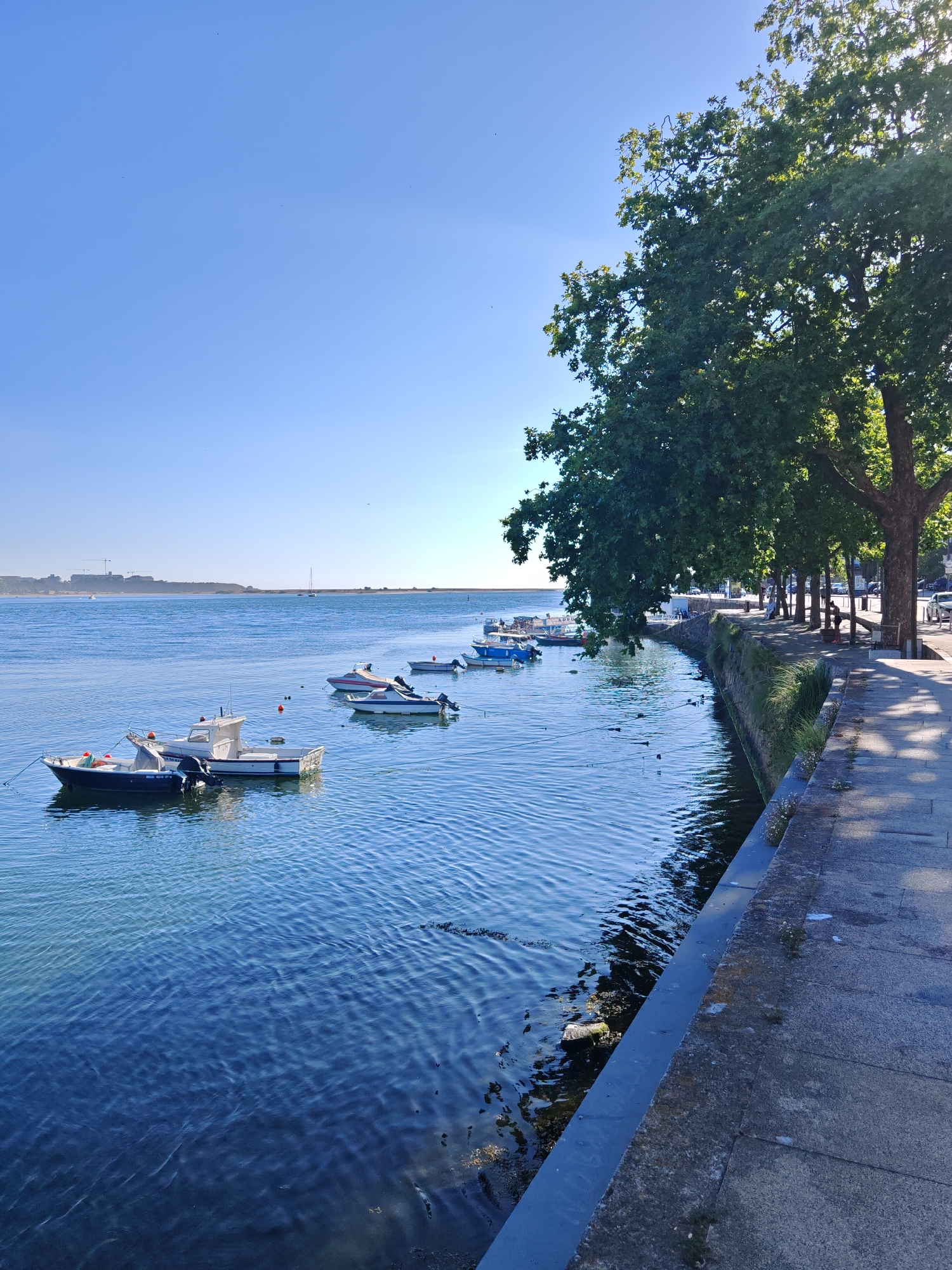 The image depicts a waterfront scene in Unknown, Unknown, on a clear day with a bright blue sky. On the left side of the frame, a body of water is visible, with several boats moored. The closest boats are a white and blue vessel and a smaller white boat, both with outboard motors. Further in the distance, more boats are scattered across the water.
The right side of the image features a paved walkway or promenade running alongside the water. This area is lined with trees, casting shadows onto the pavement. Several people are visible on the walkway, appearing to be strolling or standing. Some parked vehicles are also present along the edge of the walkway. The architecture of buildings can be seen in the background on the far right. In the far distance across the water, a hazy shoreline with some structures is visible, and construction cranes suggest ongoing development. The overall atmosphere is serene and suggests a pleasant day by the water.