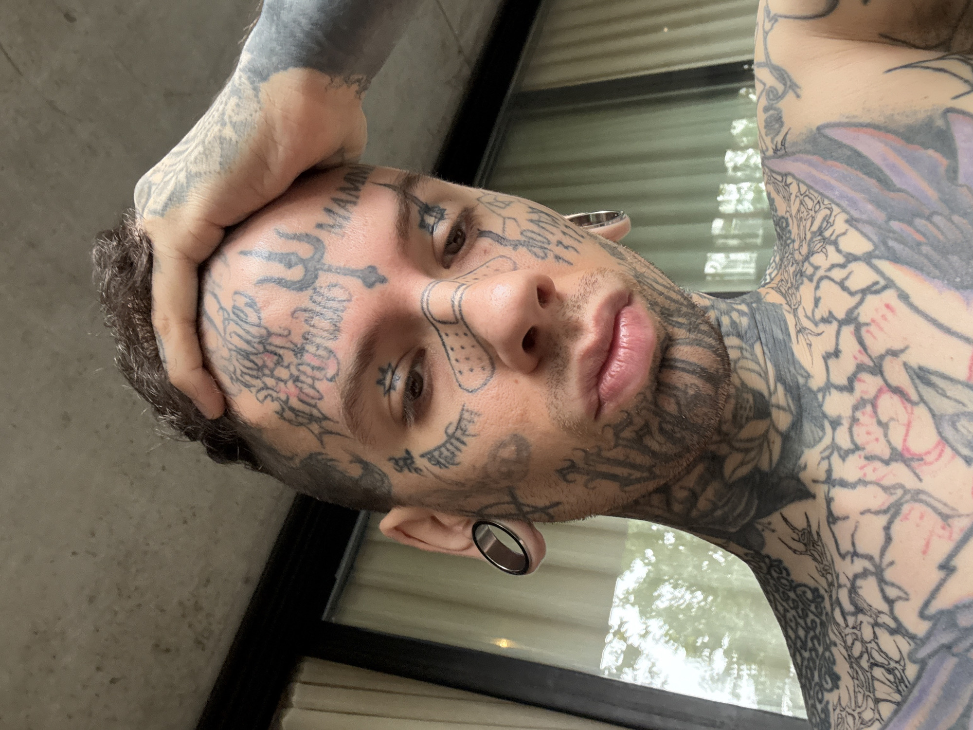 A close-up, low-angle shot shows a tattooed man with his hand on his head, facing the camera. He has extensive tattoos covering his face, neck, arms, and hand. His ears have large gauge piercings. He has short, dark hair. The background consists of an out-of-focus window and part of a wall. The location in Buenos Aires, Argentina is not deducible from the image.