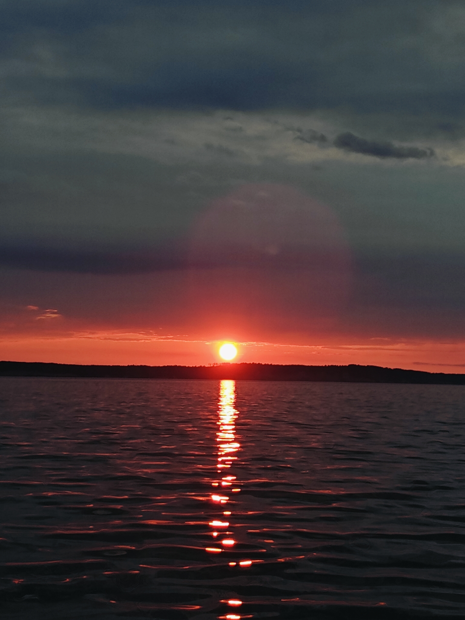 This is a vertical, medium shot photograph taken from a watercraft on the Kama River in Chaykovsky, Russia, during sunset. The sun, a bright orange orb, is low on the horizon, partially obscured by dark, heavy clouds. The sky displays a gradient of colors from deep grey at the top to vibrant reds and oranges near the horizon. A dark, silhouetted treeline forms the distant shore. The water is choppy, reflecting the fiery hues of the sunset in a shimmering path. The overall impression is one of natural beauty and tranquility.