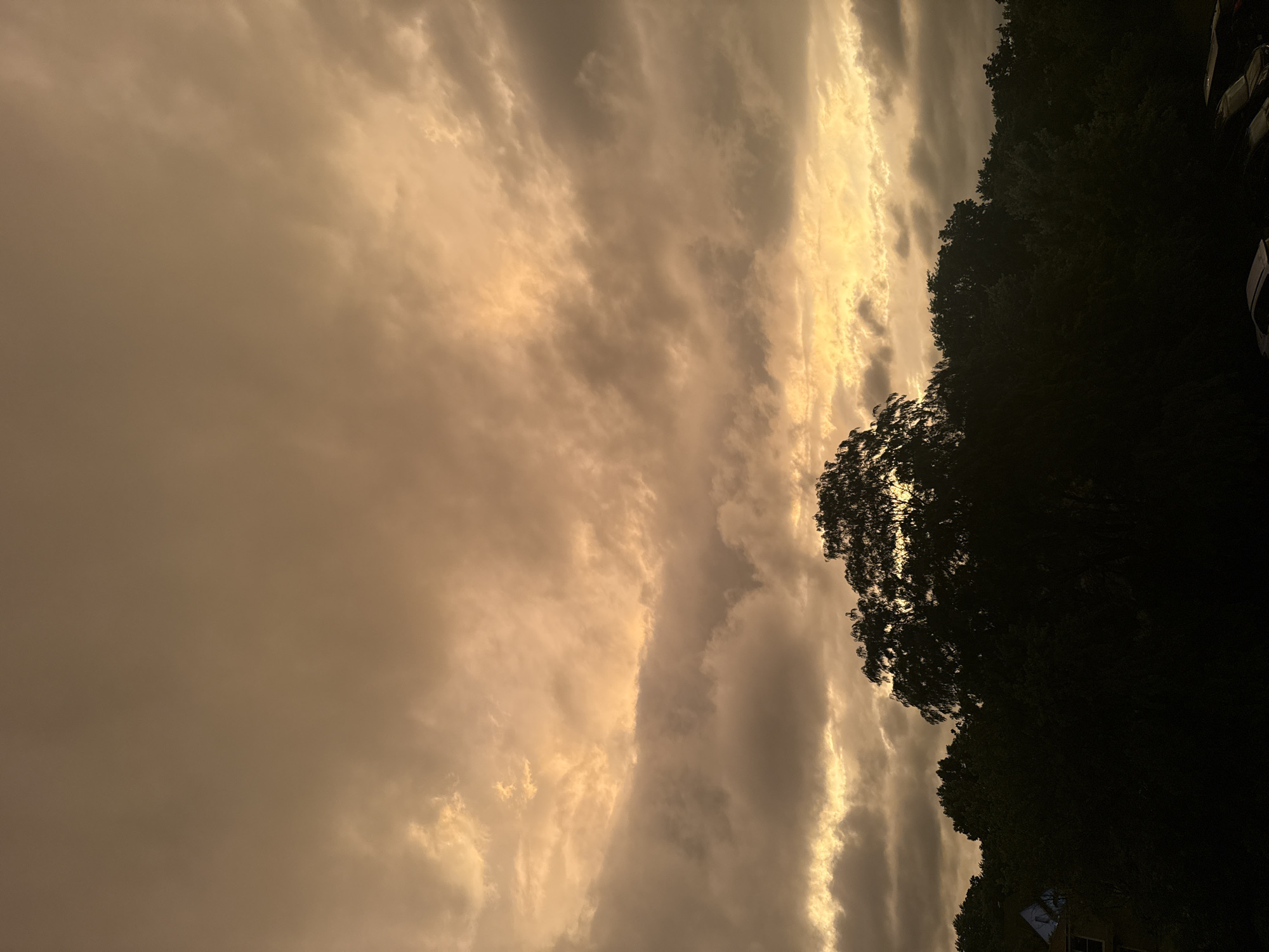 The image captures a dramatic sky over a tree line in Glendale Heights, United States of America. The sky is filled with heavy, textured clouds that are illuminated with a warm, golden light, suggesting either sunrise or sunset. The clouds are dense and layered, with some areas appearing darker and more ominous, hinting at an approaching storm.
Below the sky, a dense silhouette of trees dominates the lower half of the frame. The foliage is dark and appears lush, forming an impenetrable barrier against the light. In the far left of the image, a glimpse of a building with a blue roof is visible through the trees, suggesting the presence of residential or commercial structures within the area.
In the bottom right corner, the dark outlines of parked cars are visible, partially obscured by the trees and the low light conditions. These are the only man-made objects clearly discernible in the foreground. The overall atmosphere of the image is one of natural grandeur and impending change, with the intense sky acting as the focal point.