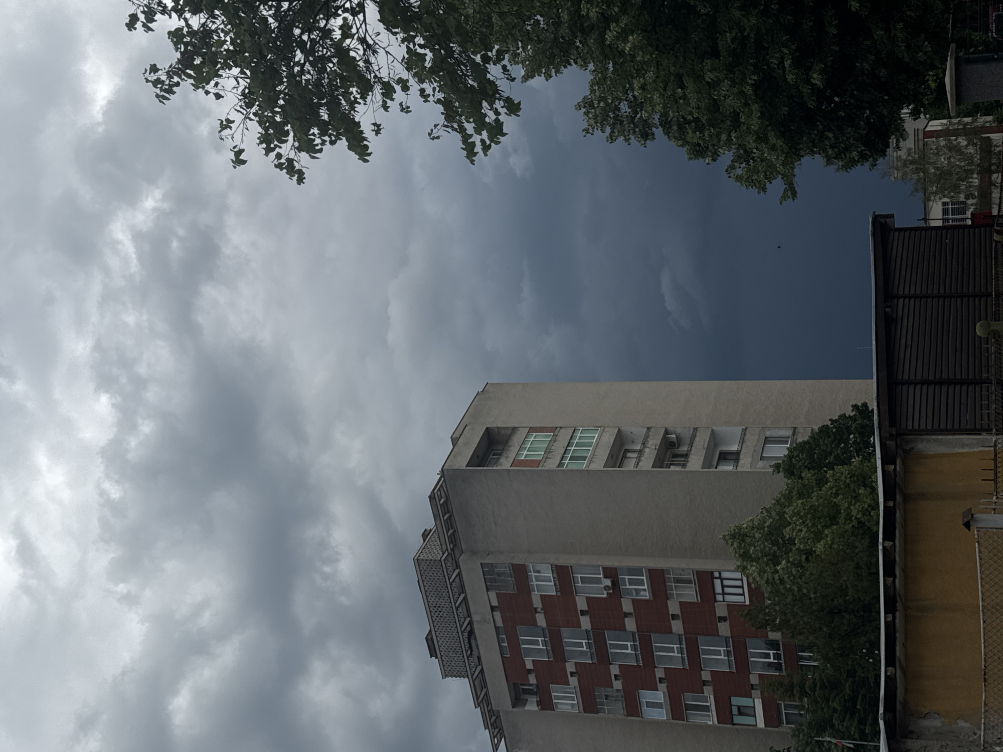 The image captures a low-angle view of a tall, multi-story building in an urban environment. The building's facade is predominantly grey with a section of red-patterned brickwork. Windows are visible across multiple floors. The sky is overcast with heavy, grey clouds. Tree branches are visible in the top corners of the image. Adjacent to the building is a fence with a yellow structure behind it. The image is slightly tilted. The location is Unknown, Unknown.