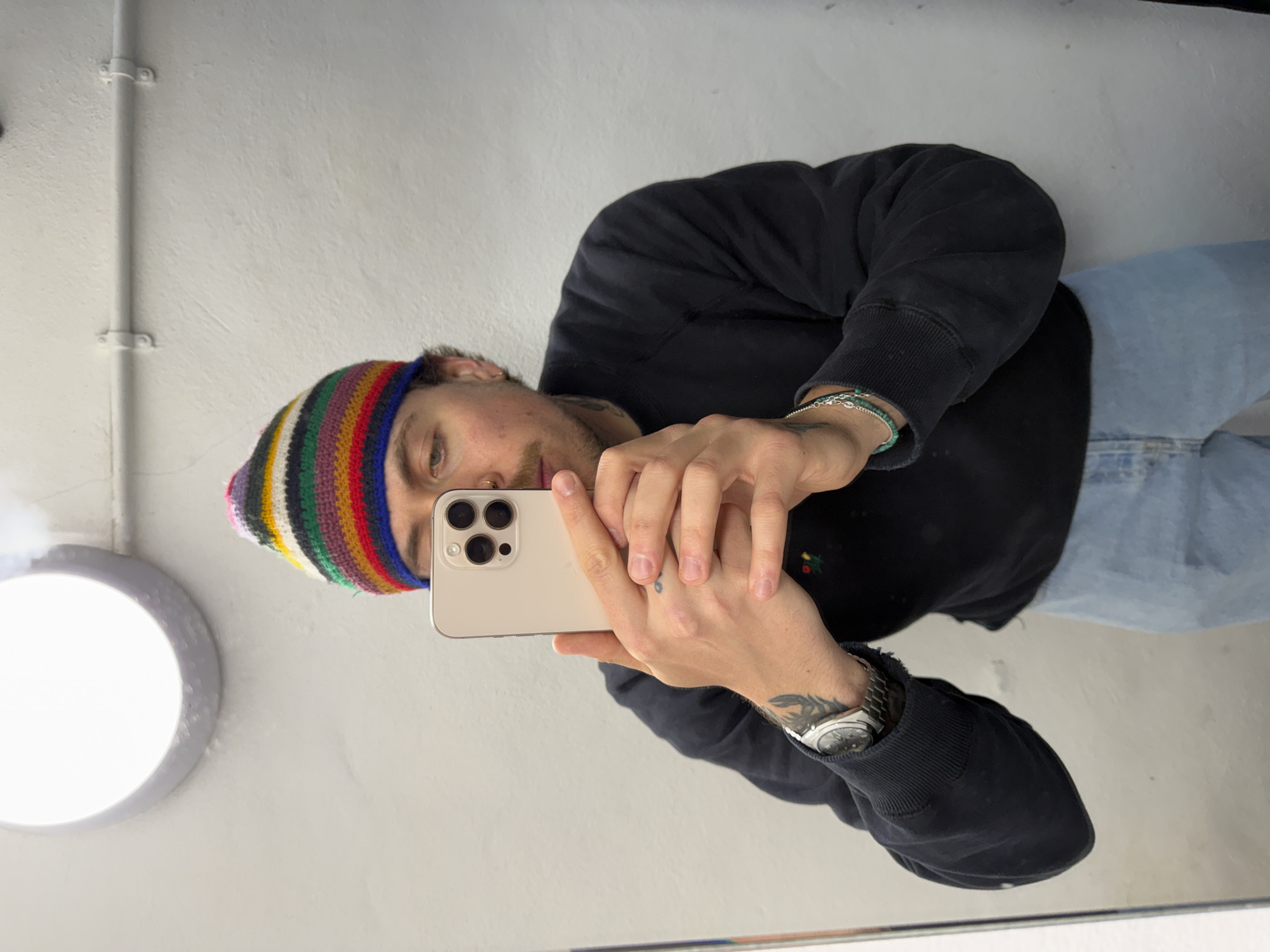 The image is a mirror selfie of a person, likely male, taken indoors. The subject is shown from the chest up, holding a light gold/beige iPhone with both hands to capture the reflection. They are wearing a dark, possibly black, long-sleeved top or hoodie, and blue jeans are visible below the waist. A colorful striped knit beanie covers their head.
The person has a neutral and focused expression while looking into the phone's screen. Notable accessories include a silver watch with a metallic strap on their left wrist, and a turquoise beaded bracelet along with a silver chain bracelet on their right wrist. Tattoos are visible on the left forearm and a small symbol is tattooed on their left index finger. They also wear a nose ring and an earring.
The background consists of a plain, light-colored wall, which appears white or off-white. White pipes or conduits run vertically along the left side of the wall, secured by clamps. A bright, circular light fixture with a grey or metallic rim is prominently visible in the lower-left portion of the image. This suggests a functional indoor environment, such as a hallway or utility area. The location context indicates this photo was taken in London, United Kingdom.
