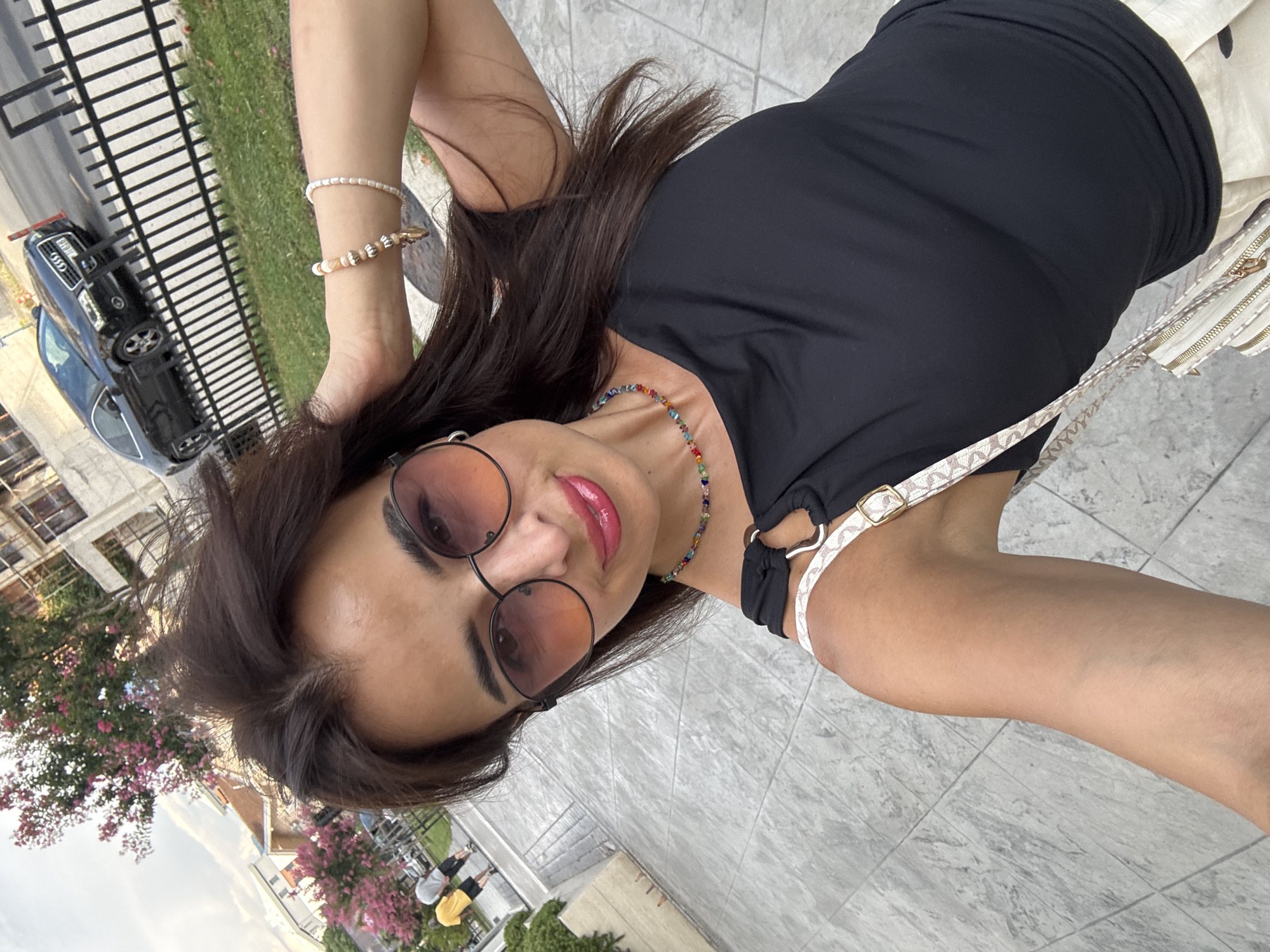 A woman with long, dark hair and sunglasses is taking a selfie outdoors. She is wearing a black top, a colorful beaded necklace, and bracelets on her wrist. The background shows a tree with pink flowers, buildings, a black car, and a black metal fence. The ground is paved with grey tiles. The woman is standing on a sidewalk.