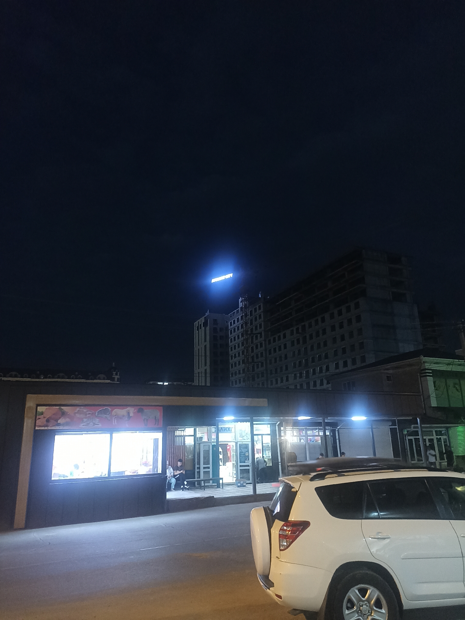 In Osh City, Kyrgyzstan, at night, a white SUV is parked on a paved street in front of a dark-colored, single-story building with illuminated windows. The building appears to be a shop or market, with a large advertisement featuring images of meat and livestock displayed above the window. Several individuals are visible inside the building and sitting on a bench outside. Further in the background, a tall, unfinished concrete building stands against the dark night sky, topped with illuminated signage that reads "MIND CITY." The scene suggests a commercial area with ongoing construction.