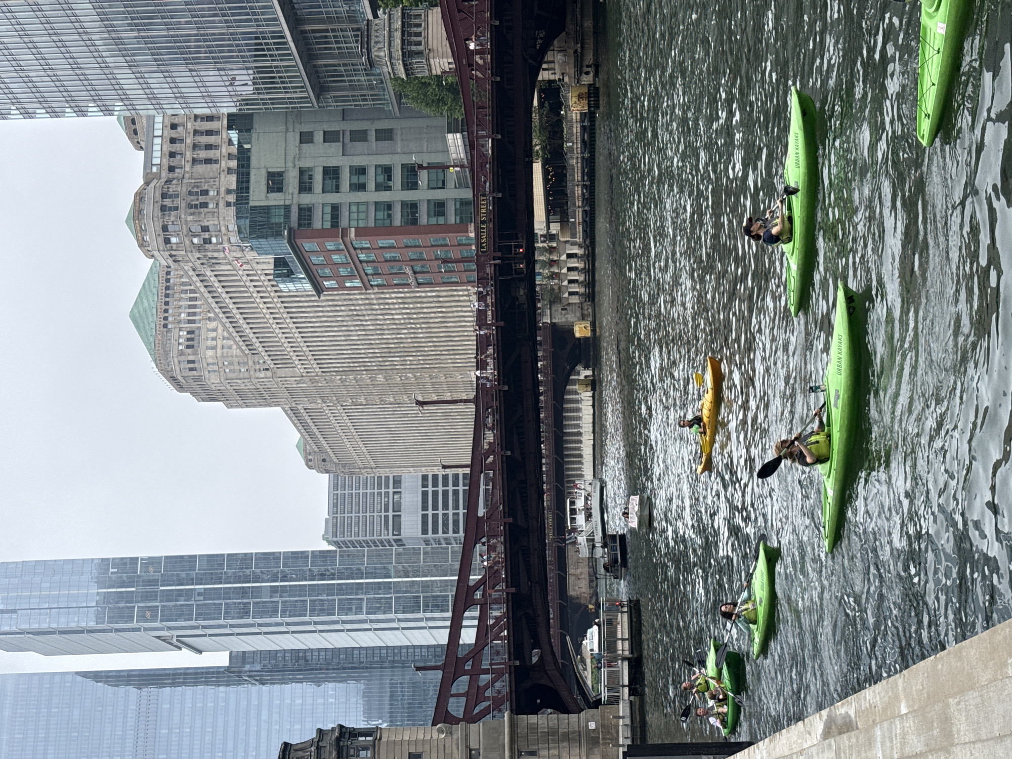 In Chicago, Illinois, USA, an overcast day finds several kayaks navigating the Chicago River. The kayaks are predominantly lime green, with some individuals sporting yellow life vests. The water is rippled and dark, reflecting the muted sky and surrounding architecture.
In the foreground, a group of kayakers moves downstream. One kayak in the very front, on the right, is occupied by a person with a dark hat, paddling with a black paddle. To their left, another lime green kayak contains a person with a light-colored helmet, also paddling. Further left, a line of three lime green kayaks are occupied by individuals paddling in unison. The furthest kayak in this line is occupied by a person with dark hair wearing a light-colored top.
Behind the kayaks, the Chicago River flows under the LaSalle Street Bridge, a red steel structure with visible pedestrian traffic on its upper level. The bridge's shadow falls onto the water below. Several boats, including a white tour boat with passengers and smaller motorboats, are visible in the middle ground, positioned further upriver.
The background is dominated by the imposing architecture of downtown Chicago. Modern glass skyscrapers with geometric facades stand tall, juxtaposed with older, ornate stone buildings featuring green roofs. The overall scene captures a common urban activity taking place amidst significant city landmarks.