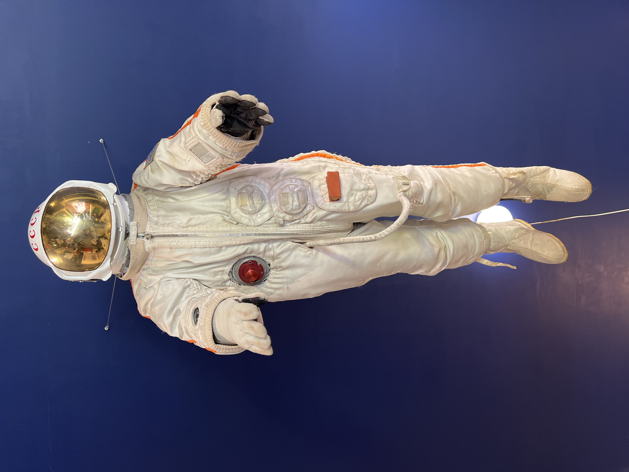 The image depicts an astronaut suit on display against a dark blue background. The suit is primarily white with orange accents on the arms and legs. The helmet has a gold visor with a reflection of the surrounding room. The suit features various details such as zippers, buttons, and a red circular device on the chest. The helmet has antennas extending from the sides. The letters "CCCP" are visible on the helmet. The location is not apparent from the image.