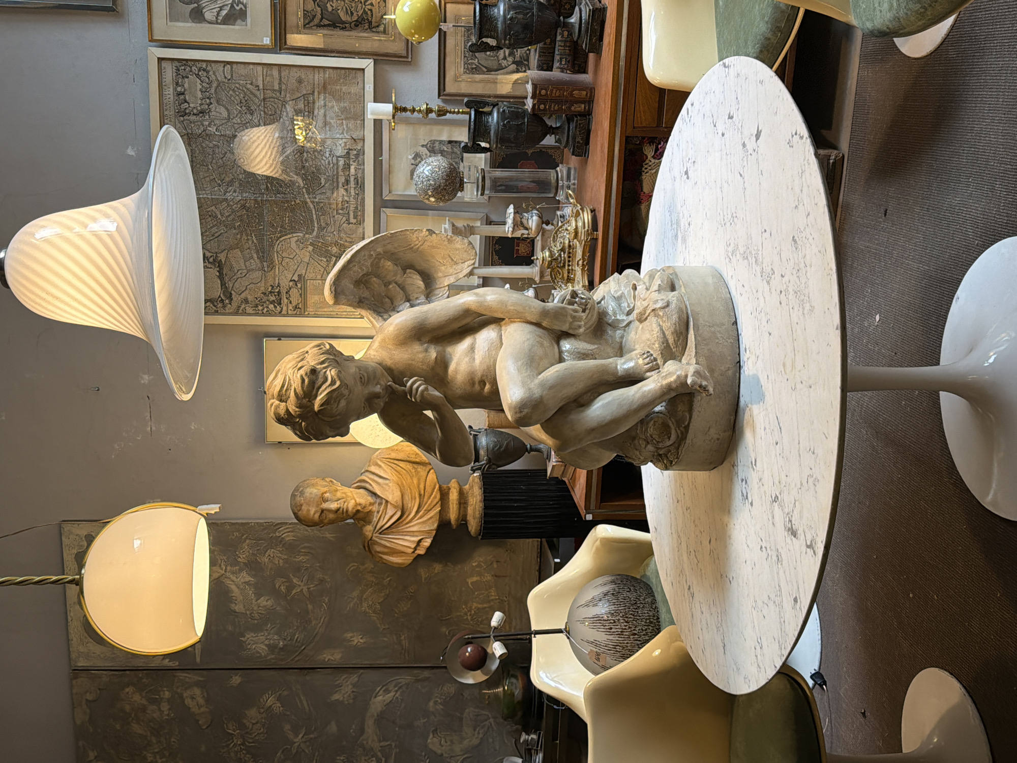 The image presents an indoor scene, likely an antique store or gallery. Several objects are prominently featured: a light-colored, textured round table with a sculpture of a nude angel on top, a variety of antique items displayed on shelves, and two different styles of floor lamps.
The items on the shelves include framed maps, vases, and decorative urns. A classical bust is also visible.
The setting appears to be in Buenos Aires, Argentina, based on the style and types of antiques typically found in that city.
