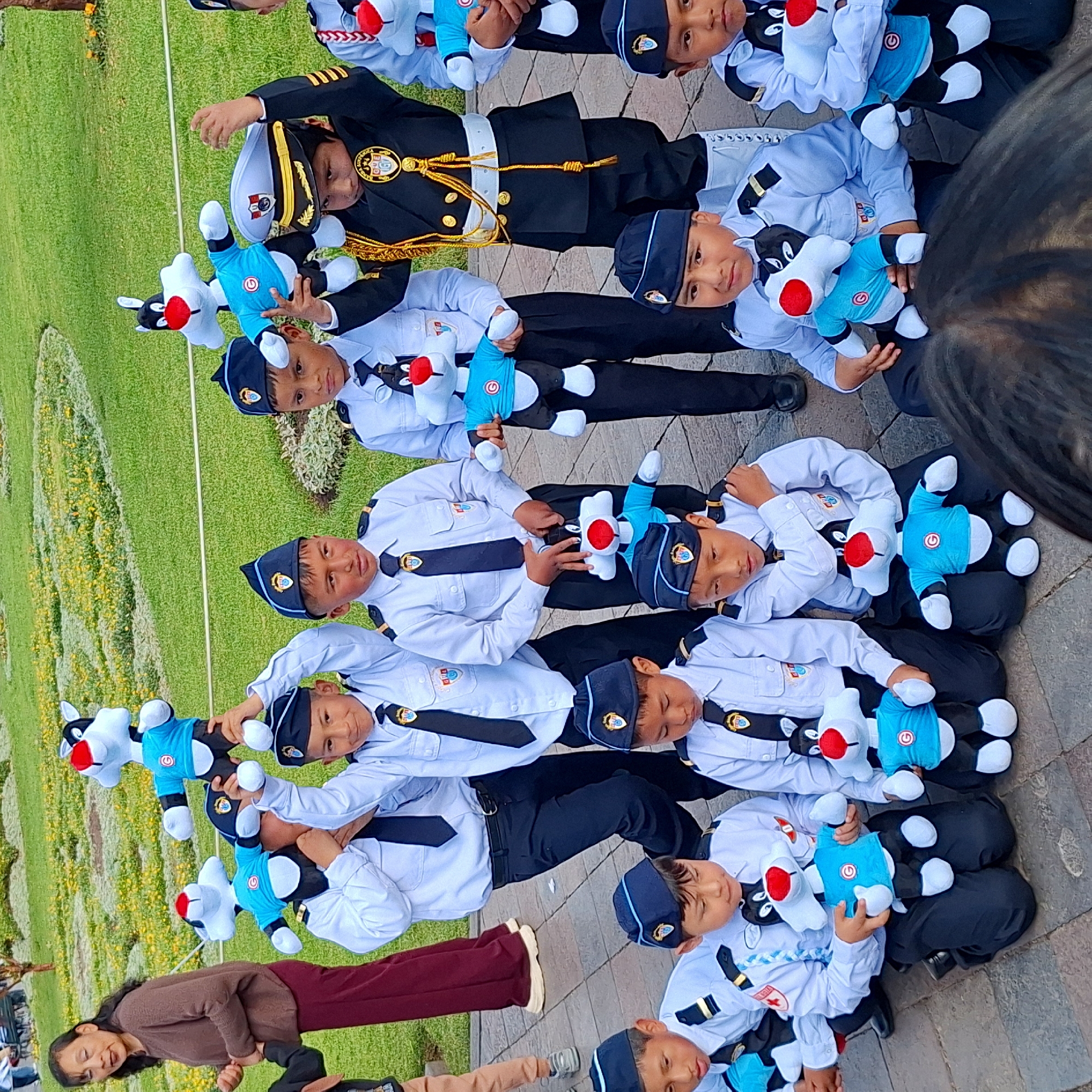 A group of children in uniform are posing for a photo outdoors in a grassy area with some yellow flowers. The children are wearing blue caps with a crest, light blue shirts with black ties and crests, and dark pants. Some of the children are holding Sylvester the Cat stuffed animals, dressed in blue shirts with a red 'G' logo. In the background, a woman in a brown sweater and maroon pants stands to the left. The scene appears to be in a park or public garden. The image composition suggests the location is the Parque de la Reserva in Cuzco, Peru, known for its gardens and historical significance.