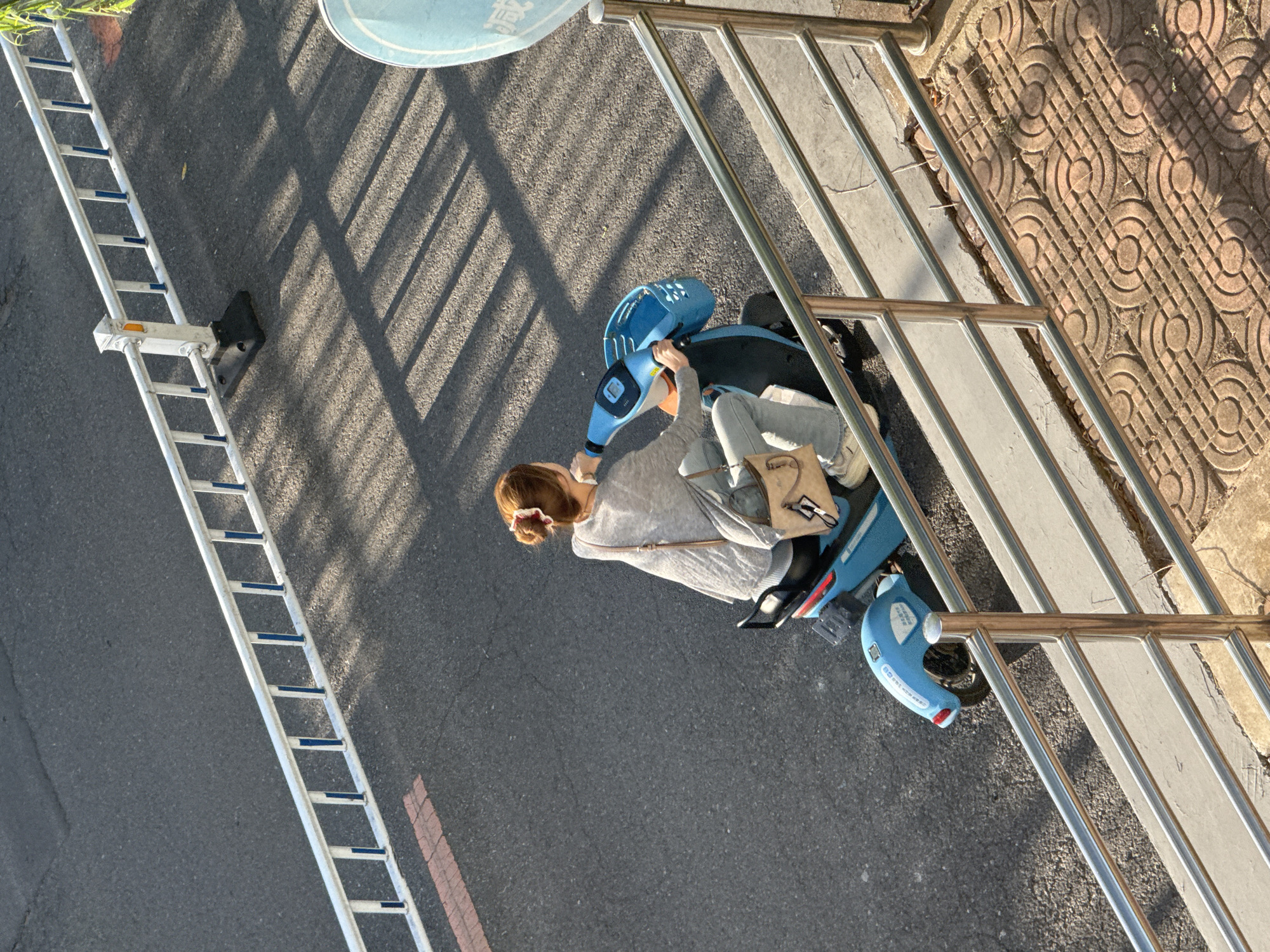 In Zhongtun, China, a woman with reddish-brown hair tied in a bun is riding a blue electric scooter on an asphalt road. She is wearing a grey long-sleeved top, blue jeans, and light-colored sneakers. A light-colored crossbody bag is slung over her shoulder. The scooter has a blue basket in front and white decals on its rear fender. The scene is viewed from above, with a metal railing in the foreground and a white fence with blue vertical accents bordering the road. Shadows from the fence are cast across the road, indicating it is daytime. The background includes a sidewalk with decorative patterned tiles and some dry vegetation.