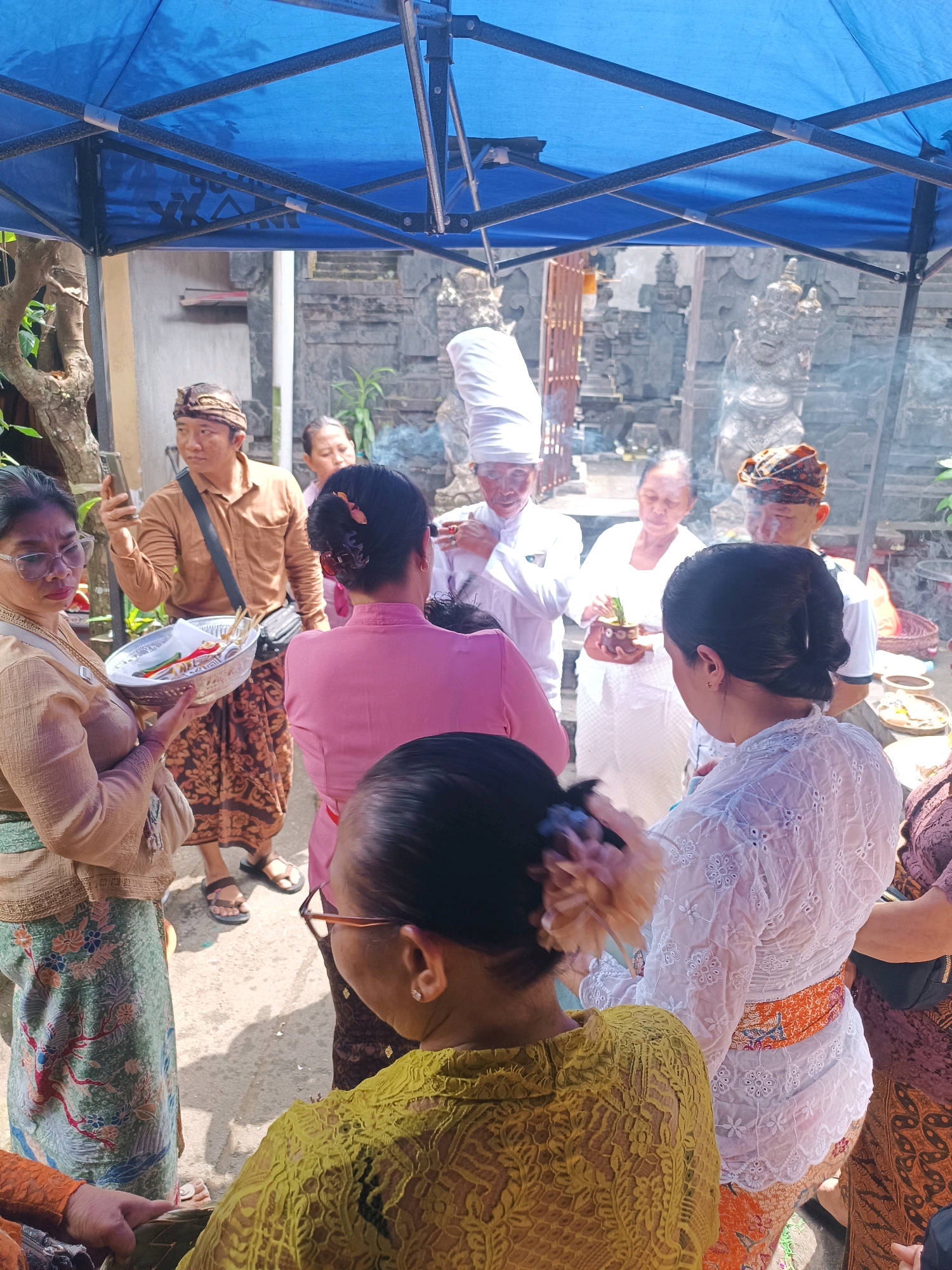 In the city of Tabanan, Indonesia, a group of people are gathered outdoors under a blue tent. The scene appears to be a religious or ceremonial event. Several individuals are dressed in traditional Balinese attire, including sarongs and ornate headwear.
In the foreground, a woman in a beige top and a patterned sarong holds a woven basket filled with various items, possibly offerings. She wears glasses and her hair is pulled back. Next to her, another woman in a pink top and a dark sarong is partially visible.
In the midground, a man in a brown collared shirt and a patterned sarong holds a mobile phone. He is looking to his left. Behind him, a woman in white holds a small container with green stems, possibly for a ritual. A man in a white headdress and robe stands next to her, with his hands clasped. Another man in traditional headwear is also present.
The background features stone carvings and architectural elements that suggest a temple or traditional Balinese compound. Smoke is visible in the air, possibly from incense or offerings. The ground is paved with asphalt or concrete. The overall atmosphere is one of communal gathering and ritual observance.
