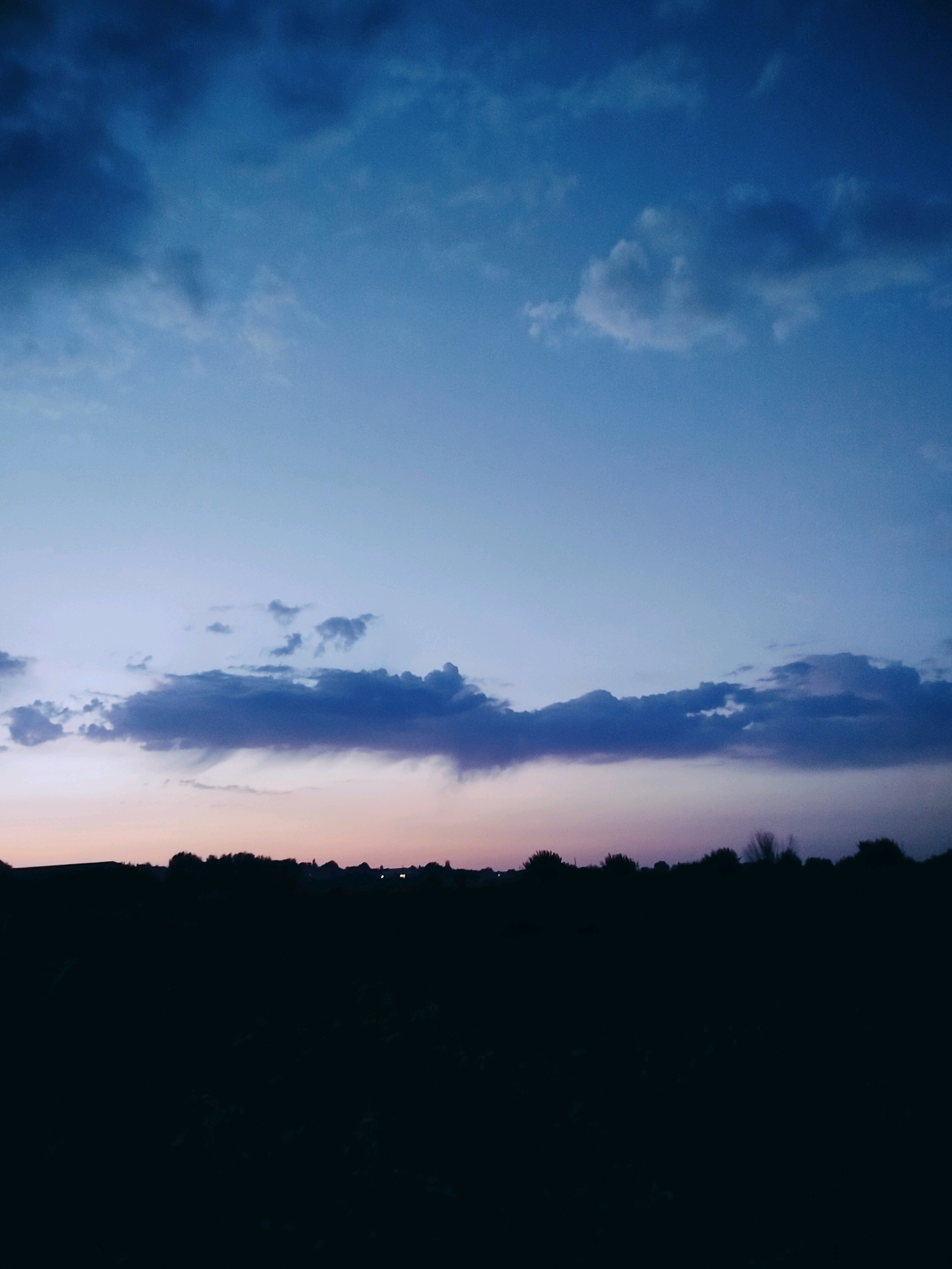 The image depicts a silhouetted landscape at dusk. A dark, undulating horizon line suggests a treeline or vegetation. Above the horizon, the sky transitions from a pale pinkish-orange near the ground to a deep blue higher up. Scattered clouds are visible in the sky, some appearing dark and heavy, while others are lighter and more wispy. The overall impression is of a rural or semi-rural setting, possibly on the outskirts of Көлкент, Kazakhstan, under a twilight sky. There are no people or visible actions in the image; it is a static landscape scene.