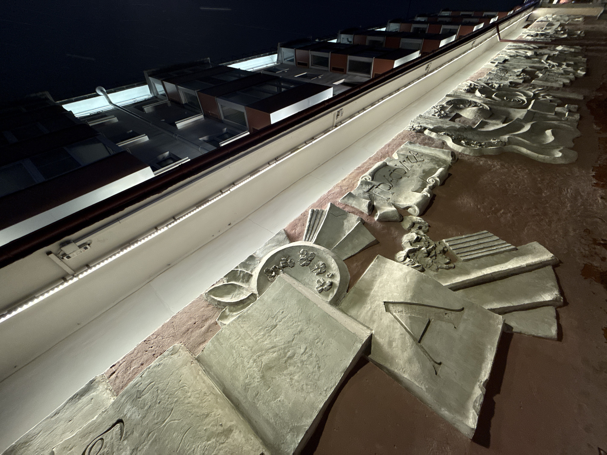 The image shows a low-angle, close-up view of a relief sculpture mounted on a building facade at night. The building, located in Perm, Russia, has a modern architectural style with multiple stories and rectangular windows. The illuminated sculpture runs horizontally along the lower section of the facade. It features a series of abstract and symbolic representations in a light-colored stone or plaster, possibly depicting elements related to education, history, or culture. Details include stylized floral motifs, book-like shapes, and inscriptions. The building is illuminated with a strip of white light running beneath the sculpture. The background is dark, indicating nighttime.