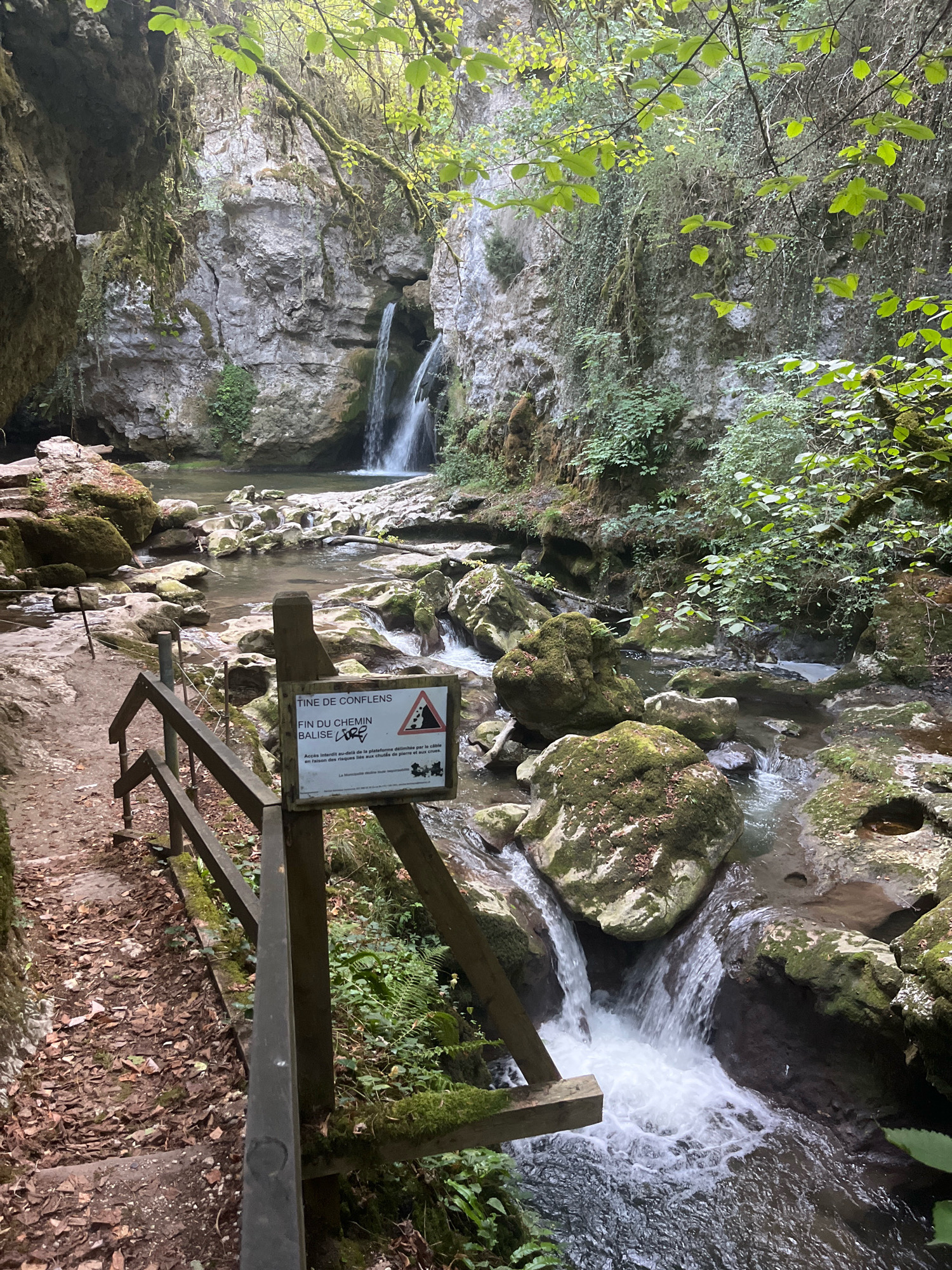 The image depicts a natural scene within the municipality of Ferreyres, Switzerland, featuring a waterfall cascading into a rocky stream. The environment is characterized by lush greenery, moss-covered rocks, and steep, rocky cliffs, suggesting a somewhat wild and untamed landscape. A wooden railing and a path lead down towards the water, indicating a designated trail for visitors. A signpost is prominently displayed near the railing, displaying text in French and symbols related to caution and danger, warning of the end of a marked path and access to a platform with risks of falling rocks and drowning. The water appears clear and is flowing rapidly over the rocks, creating a dynamic visual element. The overall impression is one of natural beauty and a sense of adventure, with the sign serving as a reminder of potential hazards.