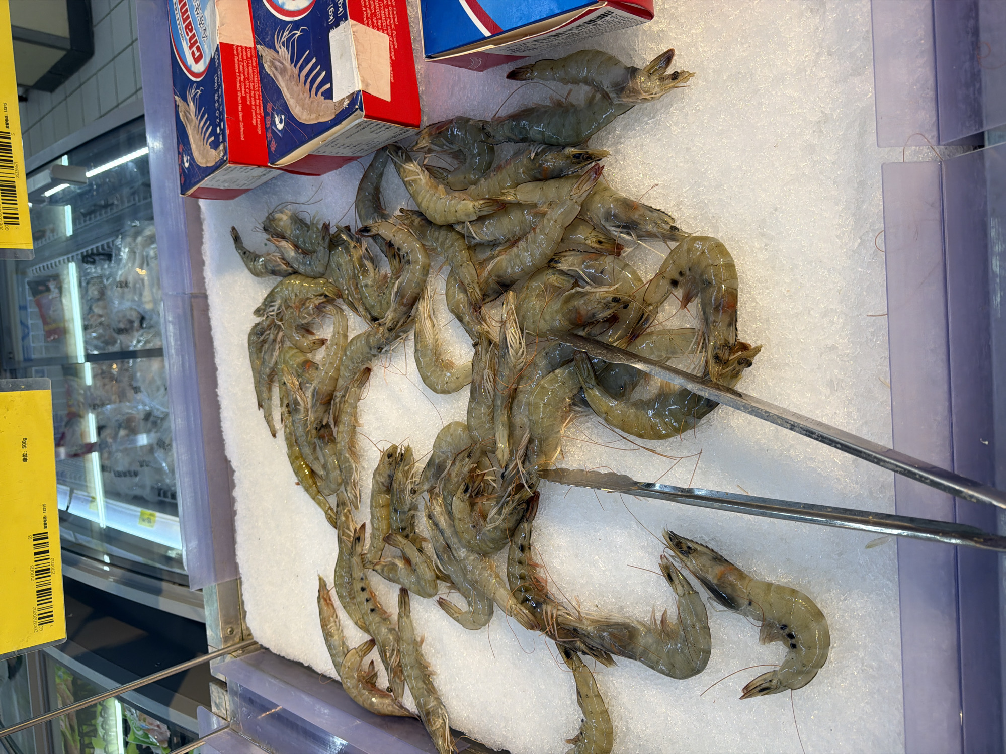 The image shows a refrigerated display of fresh shrimp at a market, presumably in Yuhuatai District, China, based on the Chinese labeling. The shrimp are piled on a bed of crushed ice in a stainless-steel tray with clear plastic dividers. A pair of silver metal tongs is also present. Boxes of Chani-branded shrimp are visible at the upper left. A yellow price tag with a barcode and Chinese text is attached to the left side of the display. The refrigeration unit behind the display contains more prepackaged goods.