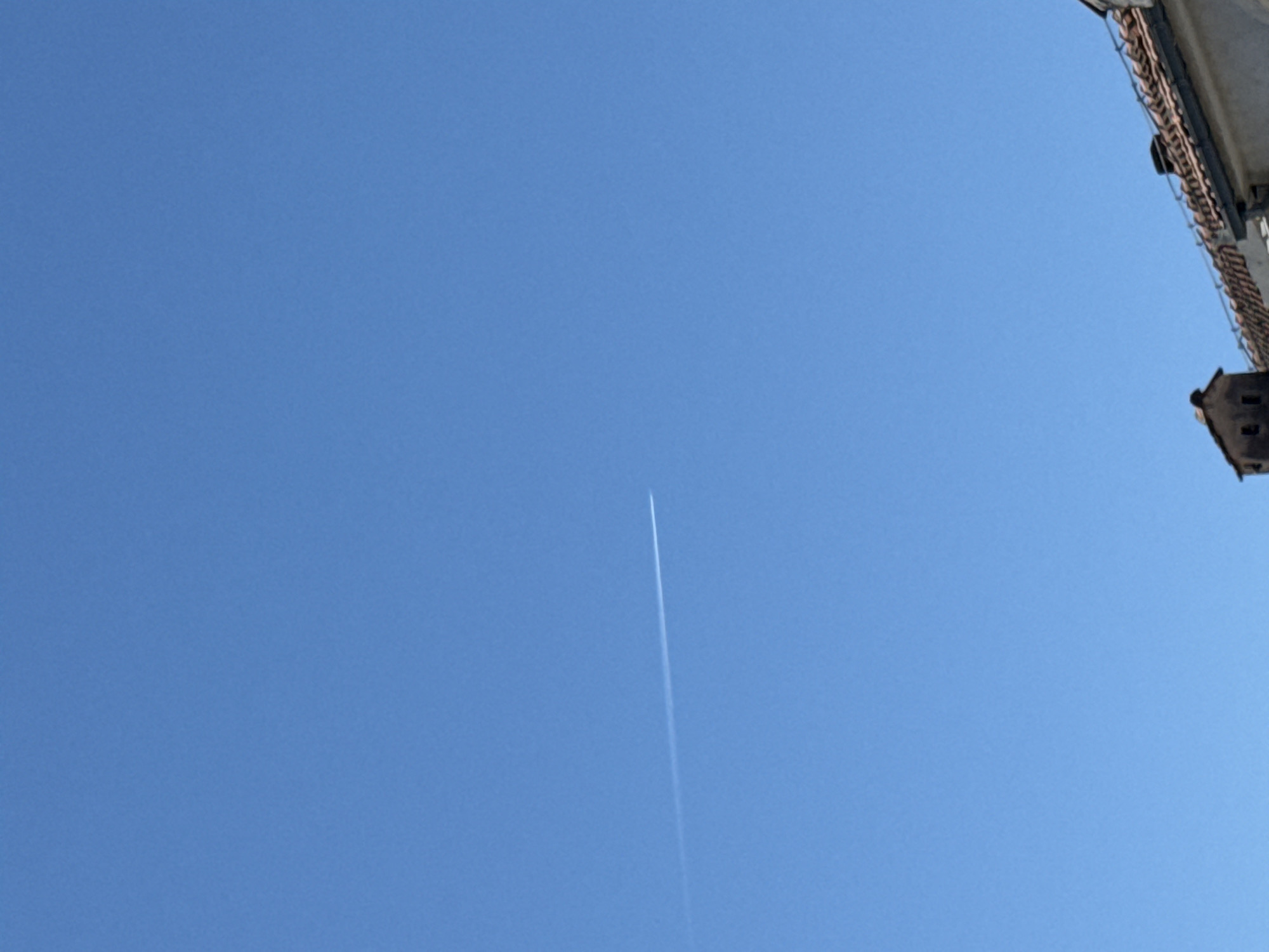 The image displays a clear, bright blue sky, indicative of a sunny day in Budva, Montenegro. A prominent, straight white contrail from an airplane diagonally crosses the central part of the frame, extending from the lower center towards the upper left. This vapor trail appears fresh and sharp against the expansive blue, suggesting a recent flight.
In the upper right corner, a portion of a building is visible, likely an upper facade or rooftop. It features distinctive terracotta roof tiles and architectural elements that suggest stone or concrete construction, consistent with Mediterranean styles. The perspective is an upward gaze, implying the photo was taken from ground level or within a built environment. The overall scene is serene, with the vastness of the sky contrasted by the human-made trace of the contrail and the glimpse of urban architecture. The bright illumination confirms it is daytime. No people or visible text are present.
