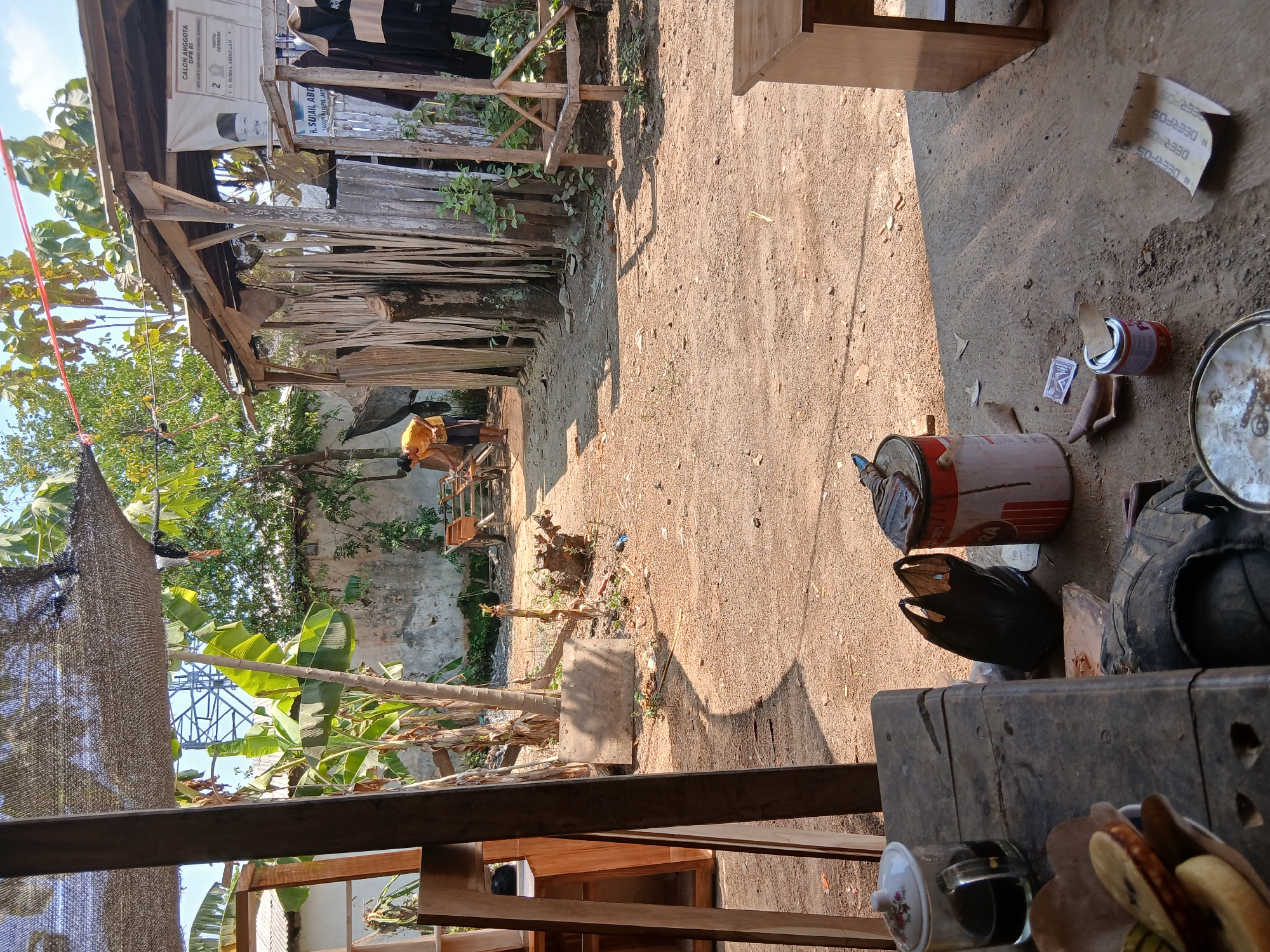 In the city of Tenggarang, Indonesia, a cluttered outdoor workshop is depicted on a sunny day. In the mid-ground, a man in a yellow shirt and dark shorts is seen working with a wooden cart. The workshop area is characterized by a dirt ground and a rustic wooden structure with a corrugated metal roof, draped with dried plant material. A poster, possibly for an election candidate, hangs on the wall of the structure. In the foreground, a weathered wooden table holds a glass of dark liquid, a bowl of sliced fruit, and a black plastic bag. Nearby, several paint cans, a brush, and other assorted items are scattered on the ground. The background features banana trees and lush green foliage, with a partially visible power tower in the distance. The overall scene suggests a working environment with a mix of tools, materials, and personal effects.