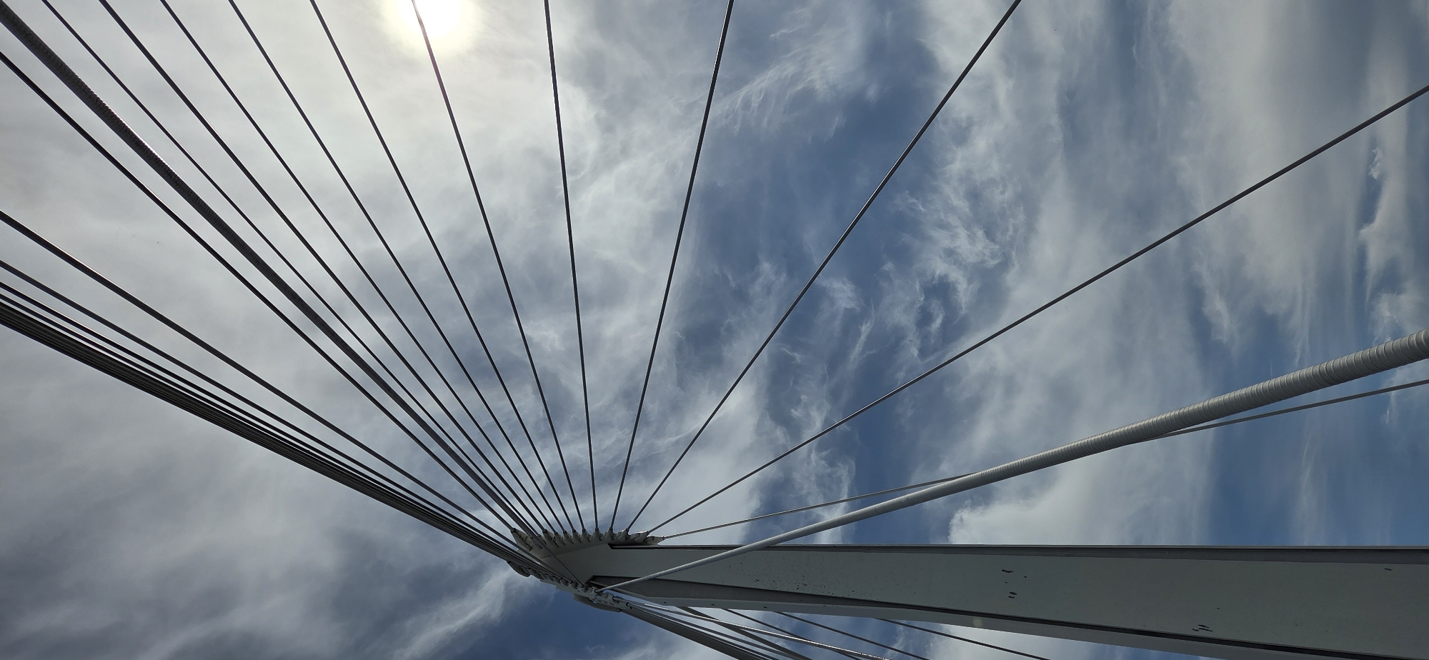 A low-angle shot captures the stark white tower and radiating steel cables of a cable-stayed bridge against a dynamic sky filled with wispy white clouds and patches of blue. The sun is partially obscured by clouds, casting a bright glow in the upper right quadrant. The bridge structure is prominent on the left side of the frame, with numerous thin cables extending upwards and to the right. The scene is devoid of people or any other discernible objects or activities. This perspective focuses on the architectural design of the bridge against a natural backdrop.