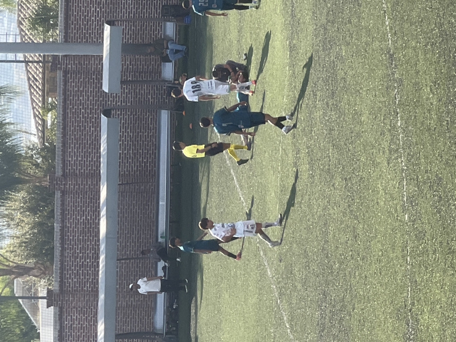 The image depicts an outdoor soccer (football) match or training session paused on an artificial turf field in León, Mexico.
The scene is dominated by a green artificial turf field marked with white lines. In the foreground and mid-ground, several individuals are present. One player, wearing a white jersey with the number "189" on the back, stands upright. Another player in a white jersey with the number "15" on the front walks towards the left. A referee, dressed in a yellow shirt, black shorts, and yellow socks, walks towards the right. One player, wearing a teal/dark green jersey with the number "7" on the back, walks towards the foreground. Another player in a similar teal/dark green jersey with the number "25" is partially visible on the far right.
Central to the mid-ground, a player lies prone on the ground while another individual, wearing a dark top and shorts, kneels beside them, appearing to provide assistance. Two small orange cones are visible on the turf near the referee.
Along the far side of the field, there is a red brick wall. In front of the wall, a long, metal-framed dugout structure with a light-colored, curved roof provides shelter over a bench. Under the left section of the dugout, one person in a white t-shirt stands, and two others are seated on the bench. Under the right section of the dugout, one individual is seated alone, looking at a device.
Above the brick wall, green foliage, including palm trees, is visible. In the distant background, a building with a reddish-brown tiled roof can be seen. The scene is illuminated by bright daylight, casting distinct shadows of the people and structures on the field.