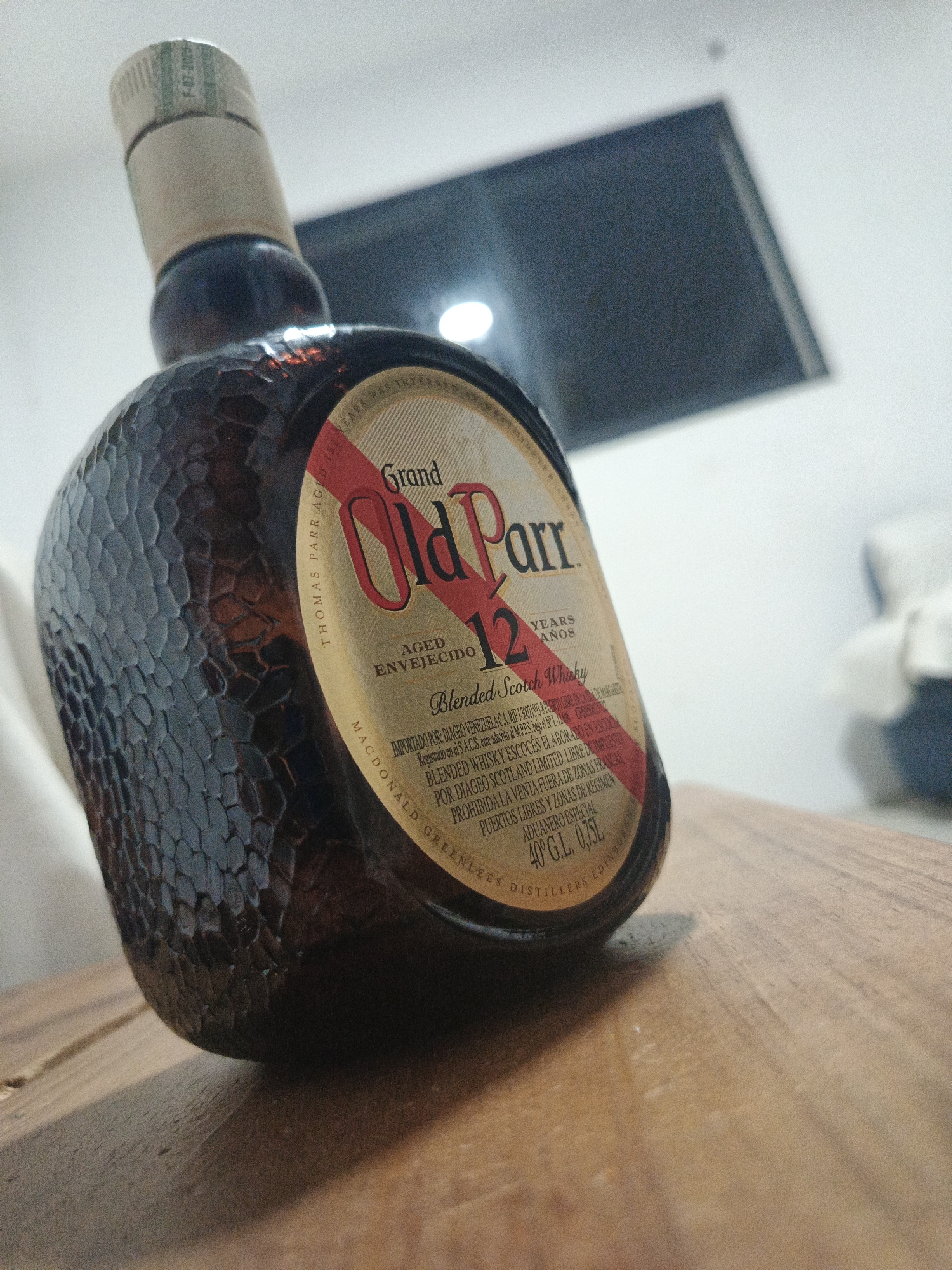 A bottle of Grand Old Parr 12 Years blended Scotch whisky sits on a wooden surface. The bottle is dark with a textured surface and has a label indicating it is aged 12 years. The label also includes information about the importer, Diageo Venezuela C.A., and states that the whisky is blended and bottled in Scotland. Text on the label also mentions "Puerto Libre de la Isla de Margarita", which is a duty-free port in Venezuela, suggesting the bottle may have been purchased there. The text "PROHIBIDA LA VENTA FUERA DE ZONAS FRANCAS PUERTOS LIBRES Y ZONAS DE REGIMEN ADUANERO ESPECIAL" indicates that the sale of this bottle is restricted to duty-free zones, free ports and special customs zones. The cap of the bottle has a printed date of F-07-2025. The bottle appears to be on a wooden table, and the background includes a blurred TV screen and some other objects. Los Robles, Venezuela, is not visibly present in the image.