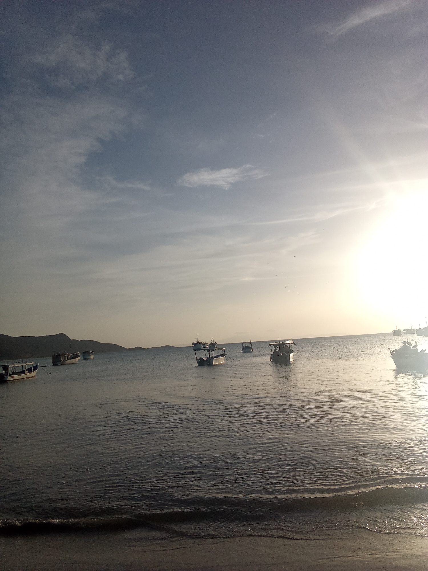 The image captures a tranquil scene at what appears to be a beach in Juan Griego, Venezuela. The sky is partly cloudy with the sun shining brightly on the right side. Several small fishing boats are scattered across the calm water. A shoreline is visible in the foreground with a wave approaching the beach. A distant mountain range is barely visible in the background.