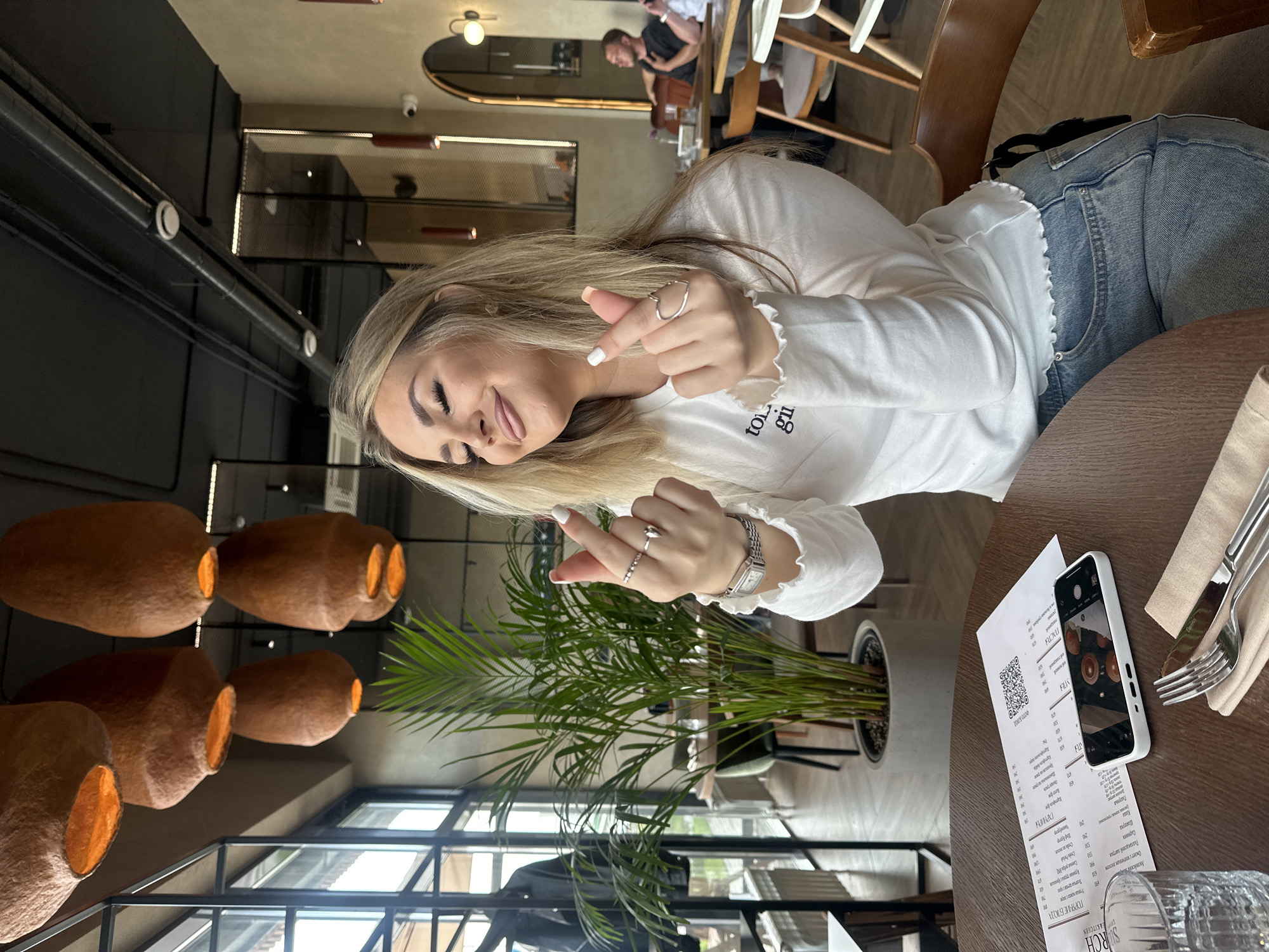 A blonde woman is seated at a dark wooden table within a modern restaurant or cafe in Усады, Russia. She wears a white long-sleeved top with ruffled edges and light blue jeans. Her eyes are closed, and she exhibits a slight smile while forming a finger heart gesture with both hands. Her right hand displays a heart-shaped ring and a thin band, while her left hand wears multiple rings and a square-faced watch. Manicured nails are visible, white on the left hand and light peach on the right. On the table, a menu with a QR code and Cyrillic text is present, alongside a white smartphone displaying an image of the restaurant's hanging lamps, a clear glass of water, and silverware resting on a beige napkin. The restaurant's interior features several reddish-brown, textured, bell-shaped hanging lamps, a black ceiling with exposed ventilation ducts, large windows on the left, and a large potted green plant. Two male patrons are visible, blurred, at a table in the background to the right, and an arched mirror with wall sconces adorns the right wall.