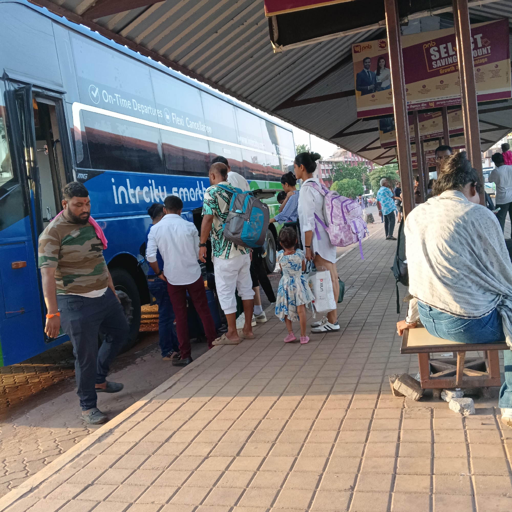 A bus stop scene in Mapusa, India, shows passengers boarding an Intercity Smart Bus labeled with "On-Time Departures" and "Flexi Cancellation." The bus is blue, and people are queued to enter. A man in a camouflage shirt stands by the door of the bus. A girl in a blue and white dress is in line, along with adults carrying bags and backpacks. Overhead, the bus stop has advertisements for PNB (Punjab National Bank). A woman is sitting on a bench to the right. The ground is paved with tiles.