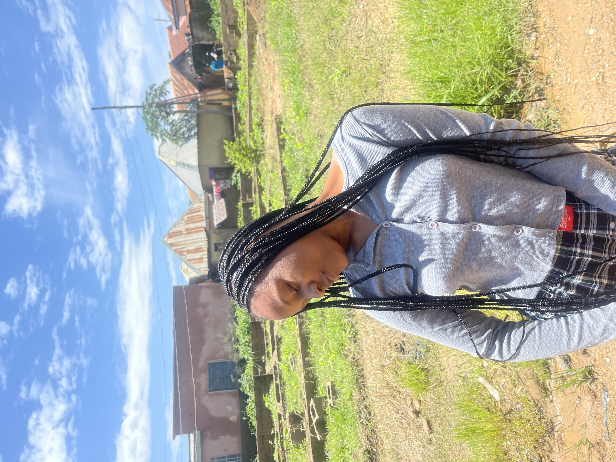 A medium shot shows a young Black woman with long, braided hair standing outdoors in Rafin Par, Nigeria. She is wearing a grey, long-sleeved, button-up shirt and plaid pants. Her eyes are closed, and her head is tilted downwards. The background features a few buildings with corrugated metal roofs, a utility pole, and abundant green vegetation under a bright blue sky with scattered white clouds. The ground is dry and dusty with some patches of grass.
