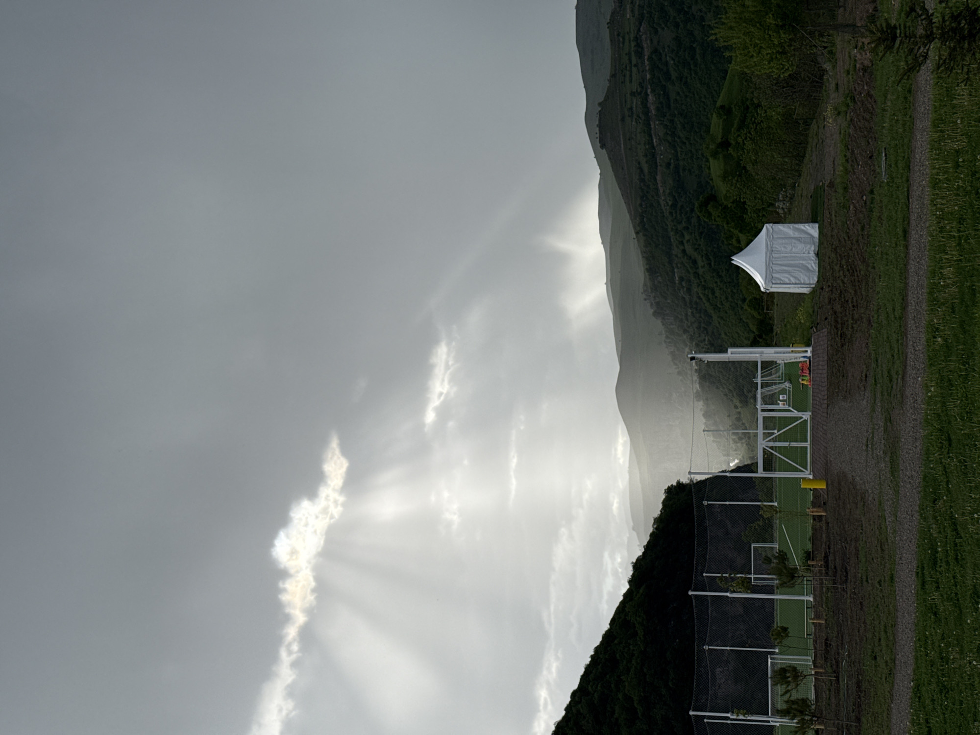 The image shows a landscape with a mountain range in the background. The sky is overcast with diffused sunlight breaking through the clouds. In the mid-ground, there's a green field, possibly a sporting area, featuring white fencing, what appears to be trees, and a small white tent. The location may be in the city of Chkalov, Armenia, but the information cannot be verified.