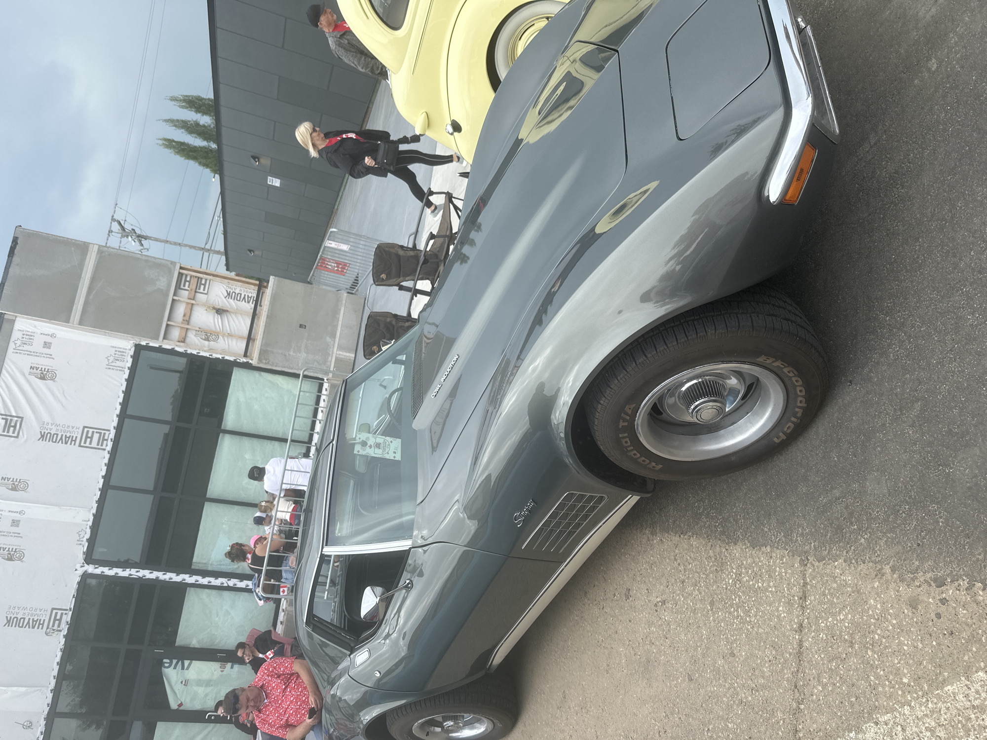 A grey Chevrolet Corvette Stingray is parked outdoors on an asphalt surface. The car is the primary subject, with its side profile prominently displayed. The iconic "Stingray" badging is visible on the car's fender. In the background, a yellow classic car, possibly a Volkswagen Beetle, is also visible. Several people are present, some standing and others seated, suggesting an outdoor gathering or car show. One person in the background is wearing a red and white patterned shirt. The setting appears to be a street or parking area adjacent to a modern building with large windows, which is partially covered with construction sheeting bearing text such as "HAYDUK LUMBER AND HARDWARE" and "TITAN." The sky is overcast, indicating a cloudy day. The time of day is difficult to ascertain precisely, but the lighting suggests it is daytime. The presence of multiple classic cars and onlookers implies an event related to car enthusiasts.