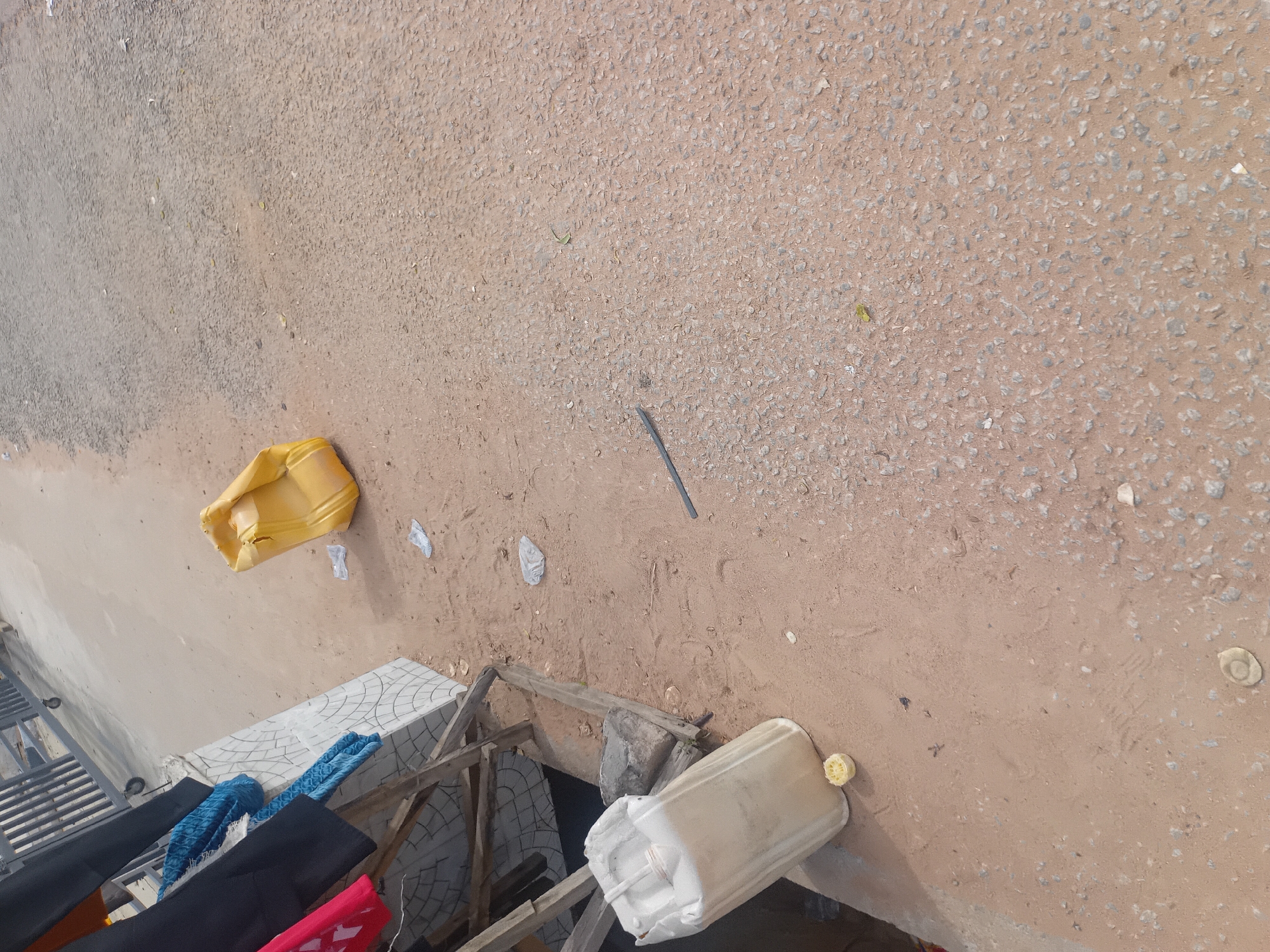 The image depicts an outdoor scene in Jalingo, Nigeria, likely a roadside or market area. In the foreground, a large, off-white plastic jerrycan with a cap sits on a sandy surface next to some wooden planks and what appears to be a tiled structure. A small, yellow, circular object, possibly a piece of fruit, lies in front of the jerrycan. To the right of the jerrycan, the ground transitions to a rough, asphalt-like surface littered with small stones and debris, including several pieces of clear plastic. Further into the background, a yellow plastic jerrycan is visible, appearing dented and possibly empty. A metal rod or pipe lies on the ground between the two jerrycans. In the upper left quadrant, a portion of a grey metal gate or fence with wheels is visible, and dark fabric, possibly clothing, is hanging on a wooden structure. Blue patterned fabric is also present, draped over the wooden structure. The environment is dusty and appears to be an unpaved or partially paved area commonly found in Nigerian cities.