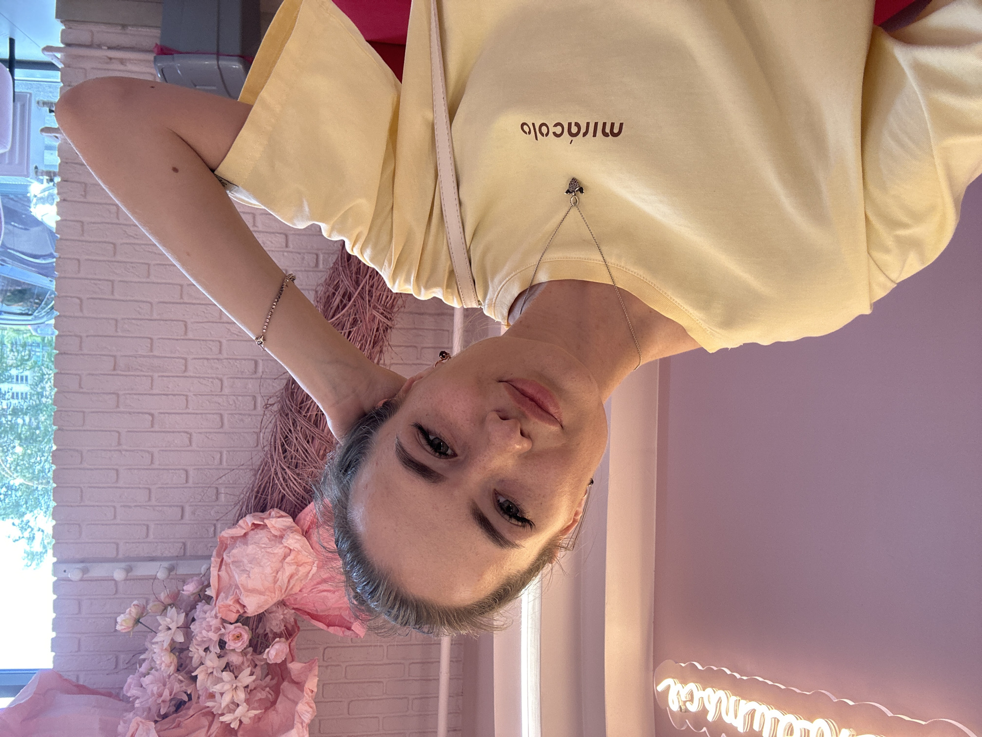 A young woman with dark hair pulled back, green eyes, and light skin is shown from the chest up, leaning her head on her right hand. She is wearing a light yellow t-shirt with "miracolo" printed in brown lettering on the left side of her chest. A silver necklace with a small dark pendant is visible. Her left arm is bent, and her hand is resting on her right arm. A light-colored strap from a bag is visible across her chest. The background is a pink brick wall with a large pink floral arrangement on the right. To the left of the woman is a lavender wall with a neon sign in white script. The lighting is bright and appears to be natural light coming from the right side of the frame. The scene is indoors, possibly in a cafe or studio.