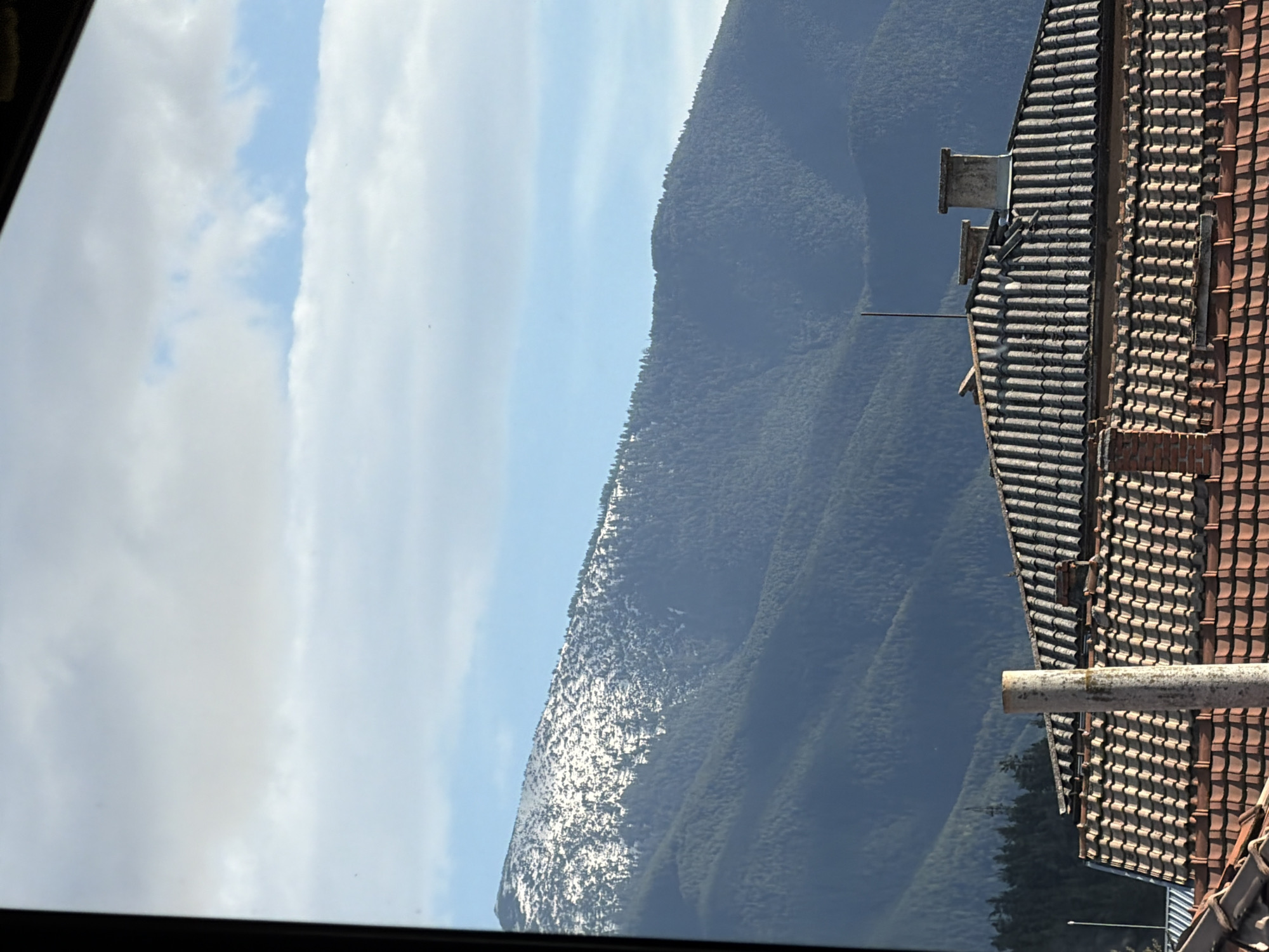 The image shows a mountain view from a window. The mountain has snow on its peak. In the foreground, there is a building with a tiled roof. The sky is partly cloudy with some blue sky visible. The location is Unknown, Unknown.