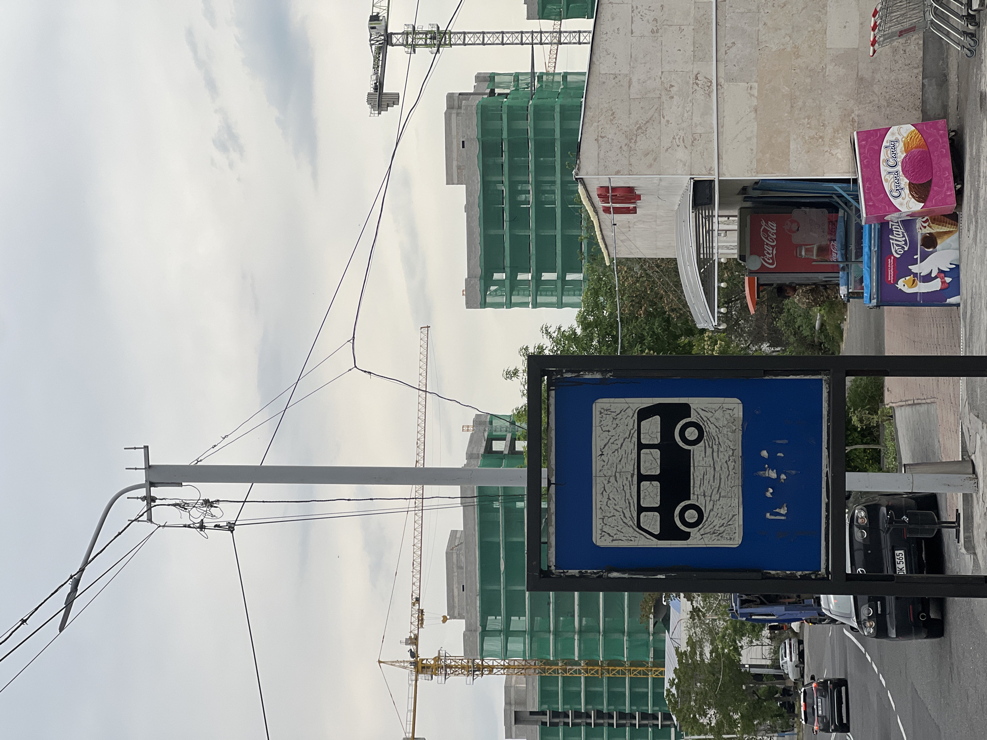 The image depicts a street scene in Yerevan, Armenia, featuring a bus stop sign prominently in the foreground. The sign is blue with a white bus icon. Behind the sign, a street with cars and a building under construction is visible. Construction cranes dot the skyline. Adjacent to the bus stop is a sidewalk with a Coca-Cola cooler and ice cream advertisements. The sky is overcast. Utility poles with electrical wires are present.