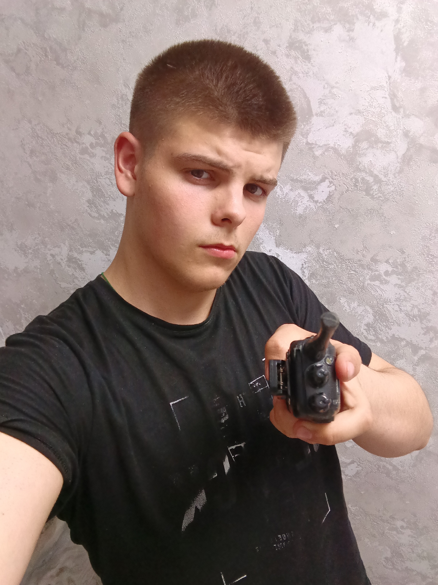 A young man with short brown hair and an intense expression holds a black walkie-talkie towards the camera. He wears a black t-shirt. The background is a textured gray wall.