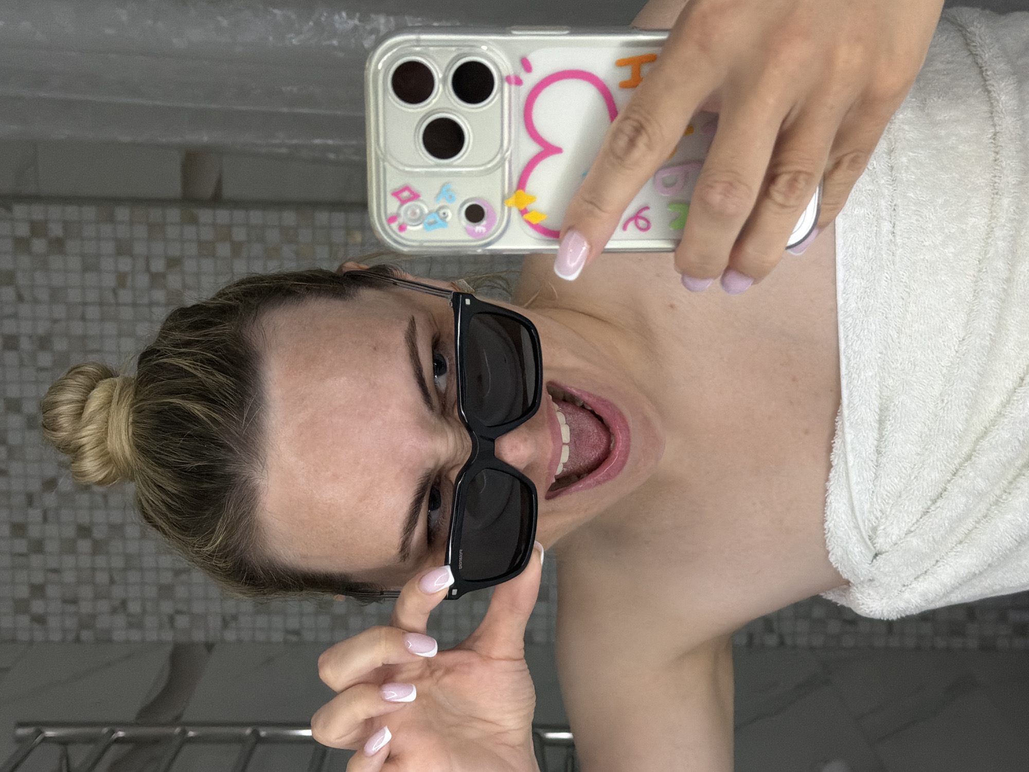 A fair-skinned woman with blonde hair pulled back in a bun is taking a selfie in a bathroom with white tiled walls. She is wearing black sunglasses and a white towel is draped over her shoulder. She is holding a phone with a clear case decorated with cartoonish images. Her nails are manicured with a French tip design and other nail color is light purple. Her mouth is open in a wide expression. The specific location within Votkinsk, Russia, is not identifiable from the image.