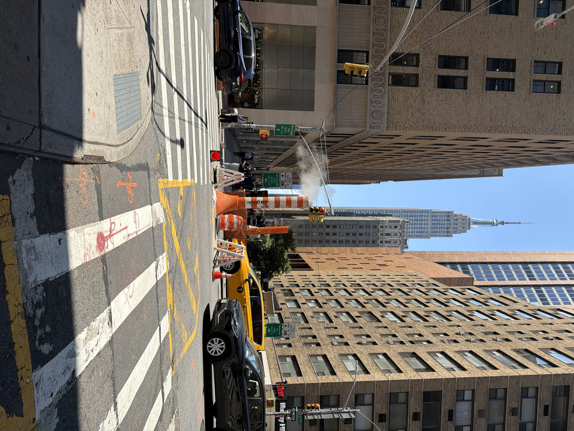 The image depicts a street scene in New York City, likely Manhattan, given the presence of the Empire State Building in the background. The orientation of the image is rotated 90 degrees counter-clockwise.
The foreground features a crosswalk with prominent white and yellow markings on the asphalt. Orange construction cones and barriers are visible, suggesting ongoing road work or traffic management. A yellow taxi cab and a dark-colored SUV are parked on the street.
In the middle ground, several buildings line the street. One building displays the text "dune" in a storefront. Traffic lights and street signs are present. A plume of steam rises from what appears to be a manhole or steam vent, adding to the urban atmosphere. Signs indicate traffic directions, with "WESTBOUND THRU TRAFFIC USE 31 St" and "LOCAL TRAFFIC PARK AVE TUN ONLY" being legible.
The background is dominated by the iconic Empire State Building, a classic skyscraper characteristic of New York's skyline. Other tall buildings with numerous windows are also visible, showcasing the dense urban architecture of the city.
The time of day appears to be daytime, with bright sunlight casting shadows. The sky is clear and blue, indicating fair weather. The overall scene captures a typical busy street corner in New York City, with a mix of vehicles, pedestrians (though not prominently featured), construction, and iconic architecture.