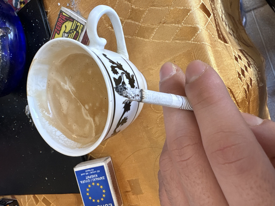 The image displays a close-up view of a hand holding a burning cigarette, alongside a cup of coffee and matchboxes, situated on a patterned tablecloth in Stefan Karadja, Bulgaria.
A light-skinned hand holds a white filter cigarette between its index and middle fingers. The cigarette exhibits a prominent column of ash and a glowing tip. Adjacent to the hand is a white ceramic cup with black decorative patterns, containing a light brown, frothy beverage identified as coffee. To the left of the cup, a blue and white matchbox is visible, featuring the European Union star circle logo and text in Bulgarian: "ZAPALKI / GYUFA KIBRIT" (Matches), confirming its origin. To the right, behind the cup, a yellow and red matchbox marked "ZEBRA" and "MATCHES" is present. A deep blue, circular glass ashtray, partially visible in the upper right background, contains scattered ash. All items rest on a golden-yellow tablecloth adorned with a repeating geometric pattern. Minor specks of ash are visible on the tablecloth and a dark, flat surface in the background.