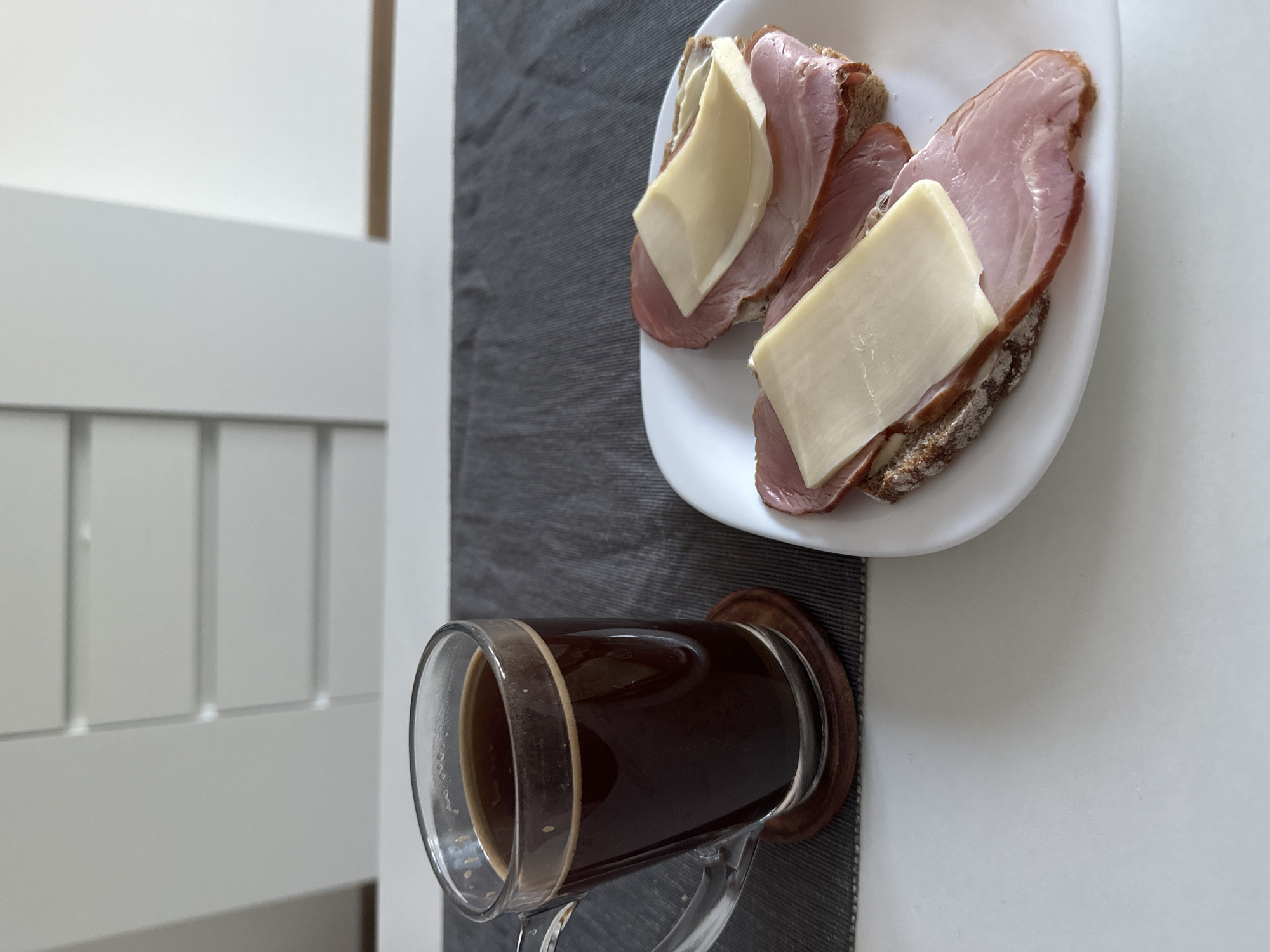 The image shows a close-up, high-angle view of a breakfast setting on a white table in Dziekanów Leśny, Poland. In the foreground on the left, a clear glass mug filled with dark coffee sits on a wooden coaster. The coffee has a light foam on top. To the right, on a white plate, are two open-faced sandwiches. Each sandwich consists of a slice of dark bread topped with several slices of ham and a slice of light yellow cheese. The background is out of focus, but appears to be a white cabinet with horizontal drawers. The lighting is bright and natural.