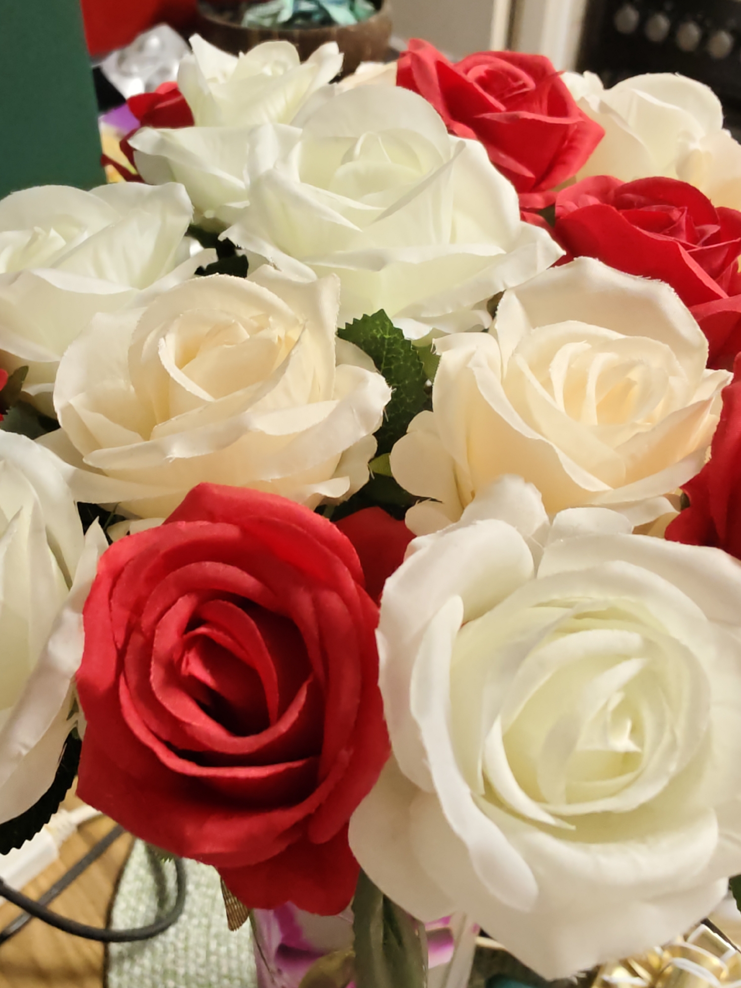 A close-up, high-angle shot showcases a vibrant bouquet of artificial roses, predominantly in shades of white, cream, and red, arranged in a vase. The roses, with their layered petals, are the central focus. Some green foliage is visible amongst the blossoms. In the background, out of focus, are hints of a domestic setting, including what appears to be kitchen appliances and other decorative items. The lighting is bright, illuminating the artificial flowers and casting soft shadows. There is no identifiable presence of Petah Tikva, Israel, or any urban environment in the image; the scene appears to be set indoors, possibly in a home.